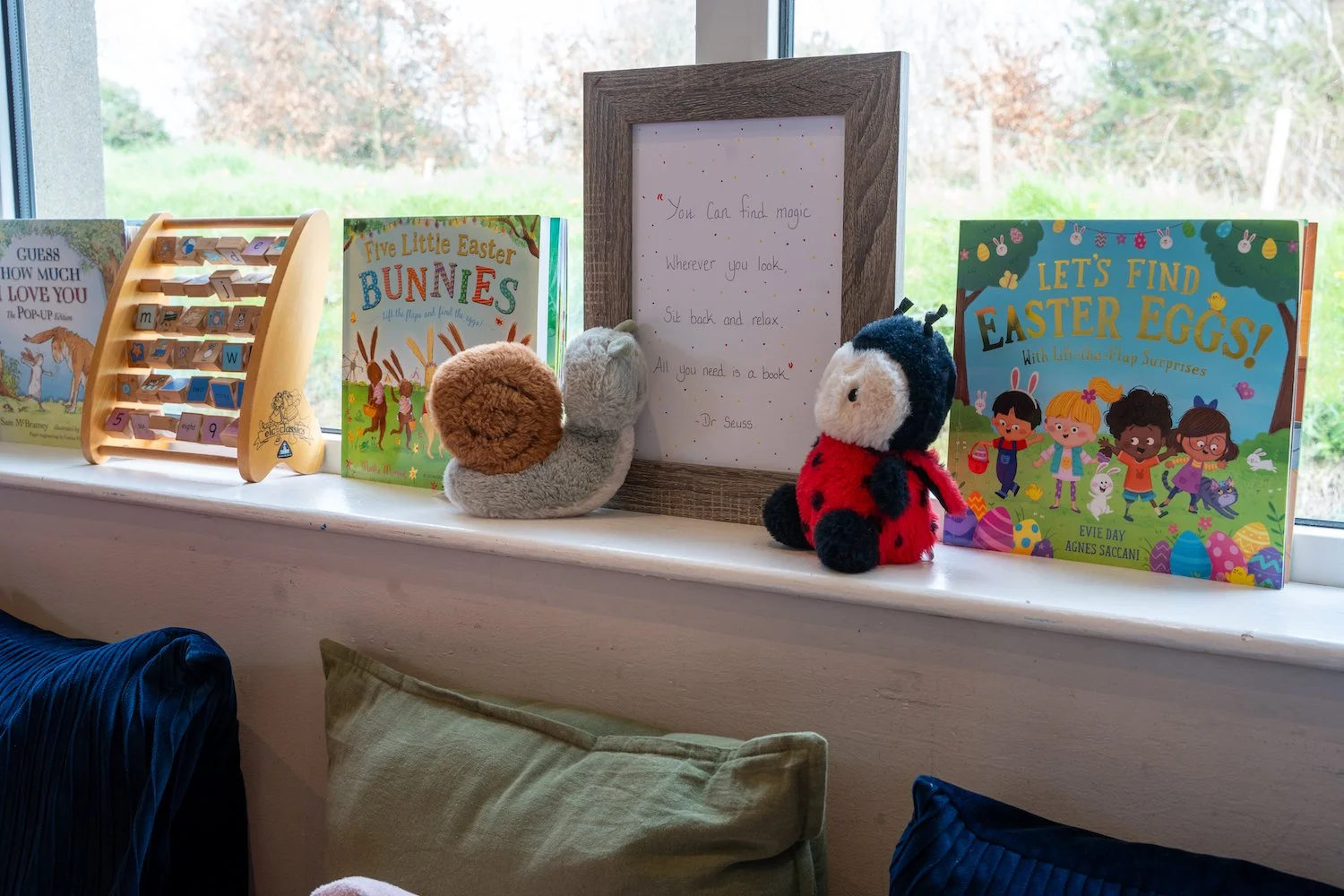 Books, plush toys, and a framed quote on a windowsill with a background of trees outside.