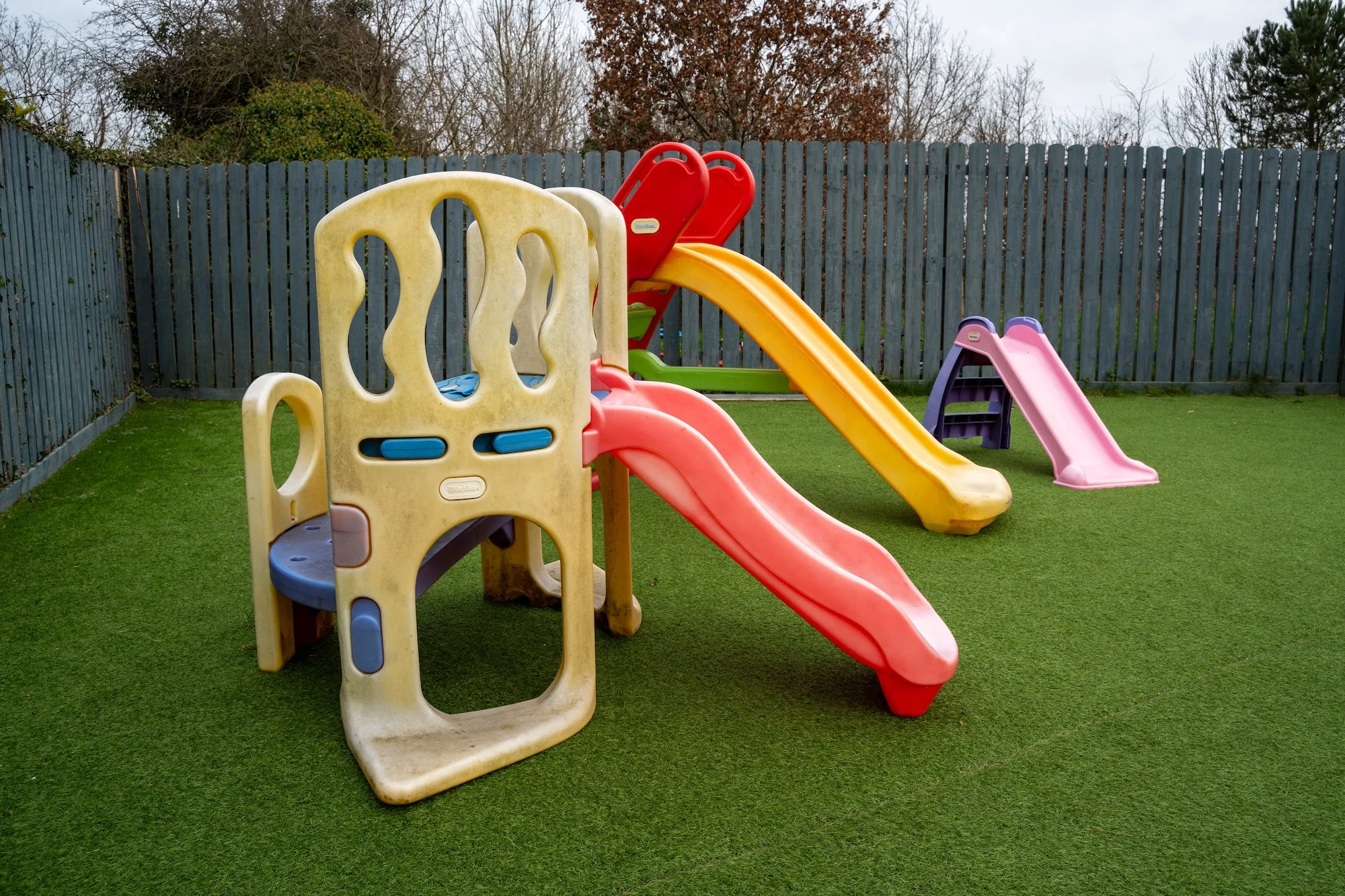 Colorful plastic playground slides in an outdoor yard enclosed by a wooden fence, with artificial grass surface.