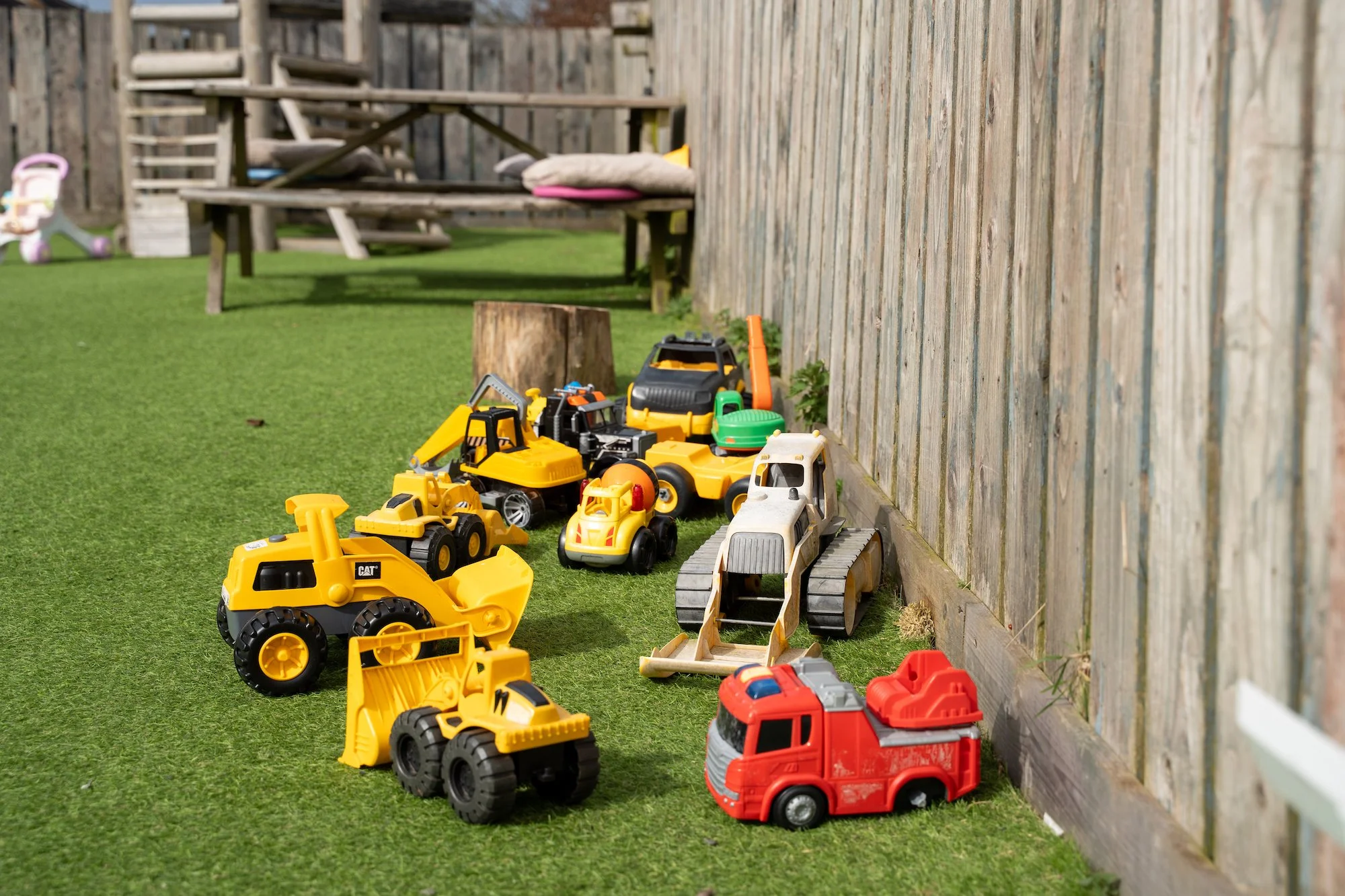 Various toy construction vehicles lined up on a grassy yard next to a wooden fence.