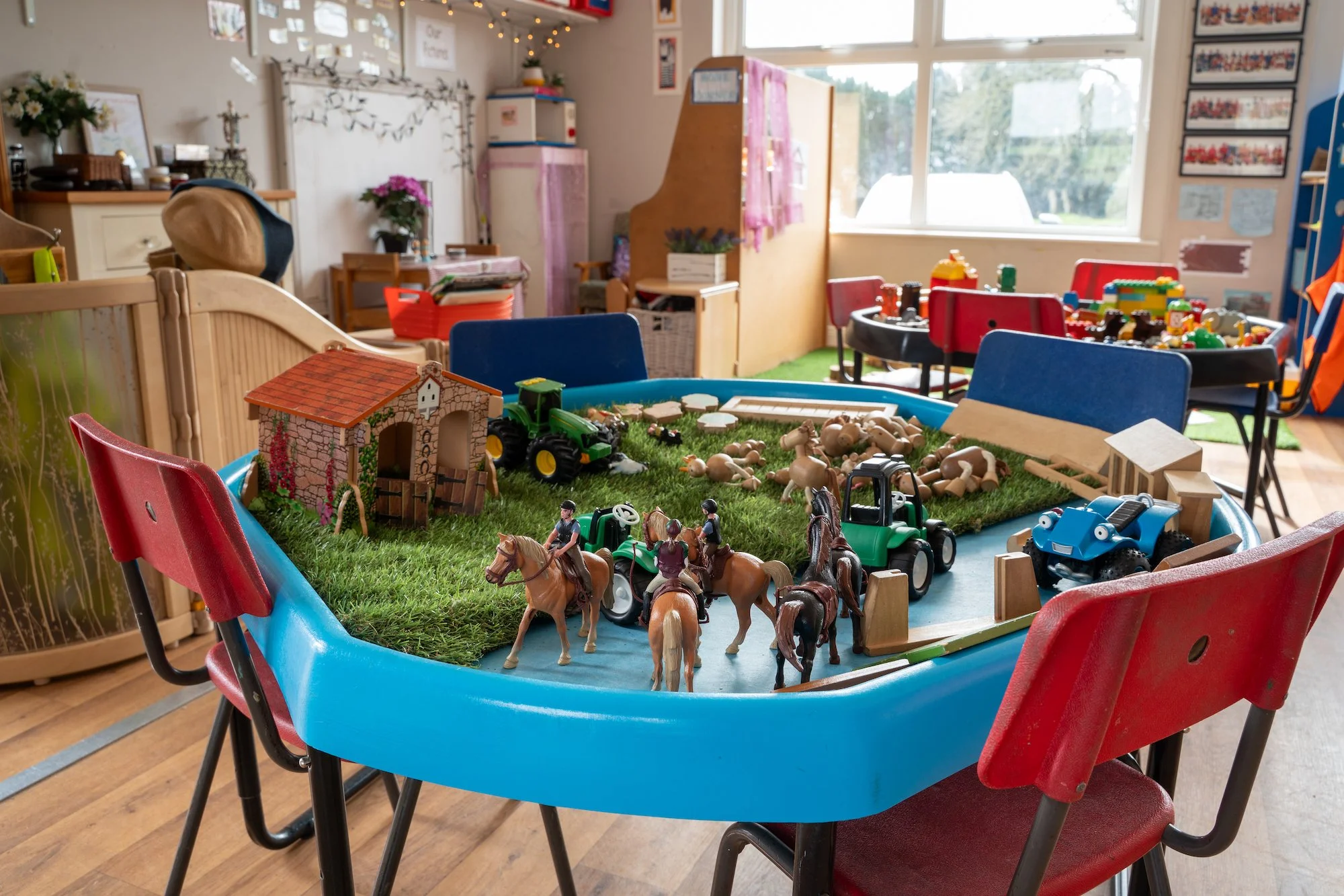 Table with toys including miniature tractors, horses, and farm animals on a grassy surface inside a classroom.