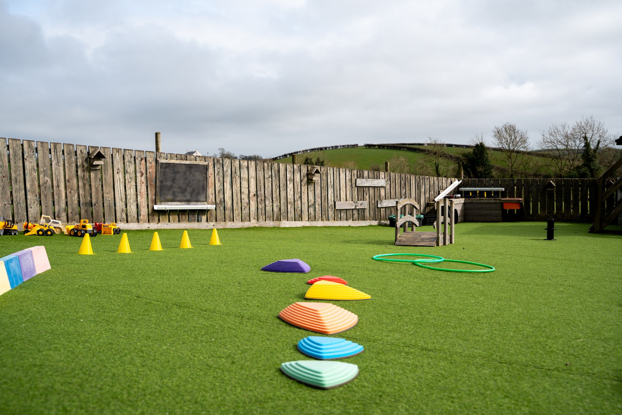 Children's outdoor playground with colorful play equipment, including cones, platforms, and a slide, on artificial green turf enclosed by a wooden fence, with a rural landscape and cloudy sky in the background.