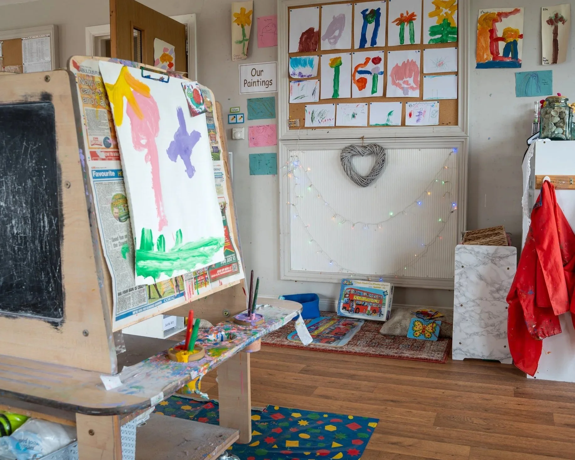 Children's art classroom with paintings on the wall, an easel with watercolors and a painting in progress, and various arts and crafts supplies.