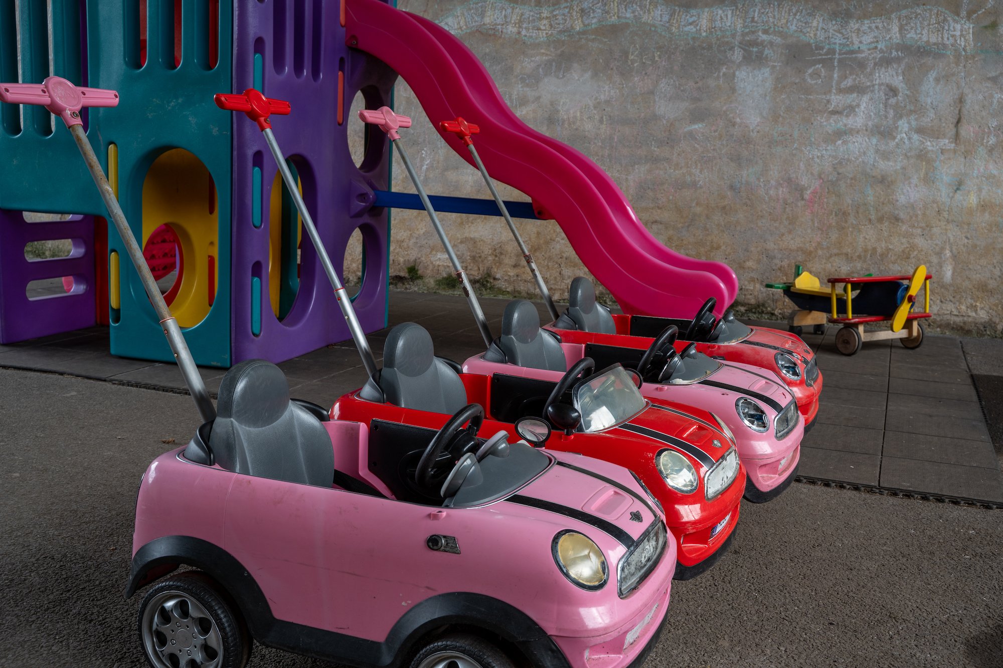 Three children’s ride-on toy cars in pink and red, parked on pavement in front of a colorful play structure with purple, blue, yellow, and pink components and a pink slide. A small yellow wagon is on the right side.