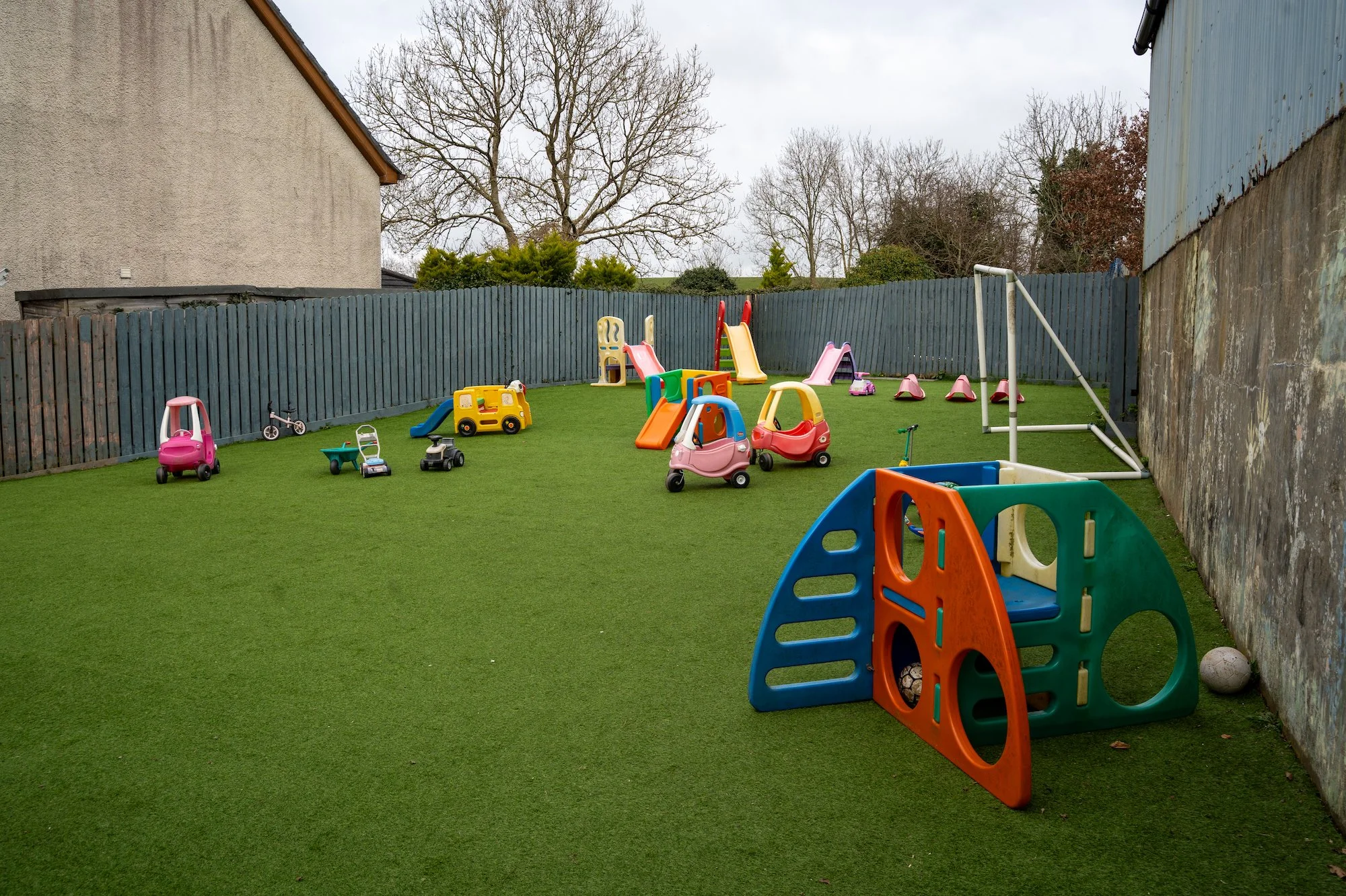 Children's outdoor playground with plastic slides, ride-on toys, a soccer goal, and a small ball on artificial grass, enclosed by a blue wooden fence and stone wall.