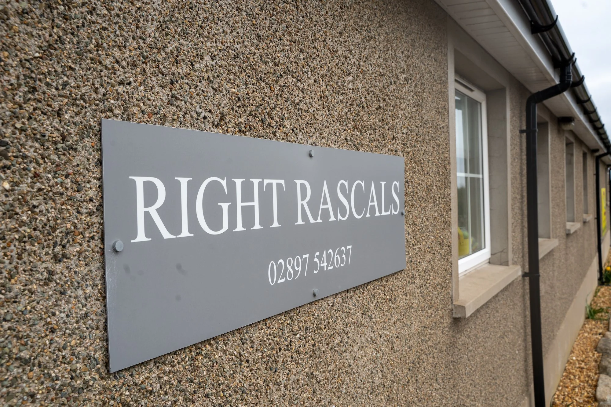 Close-up of a house exterior with a nameplate that reads 'RIGHT RASCALS' and a phone number, attached to a rough-textured wall with windows and a downspout in the background.