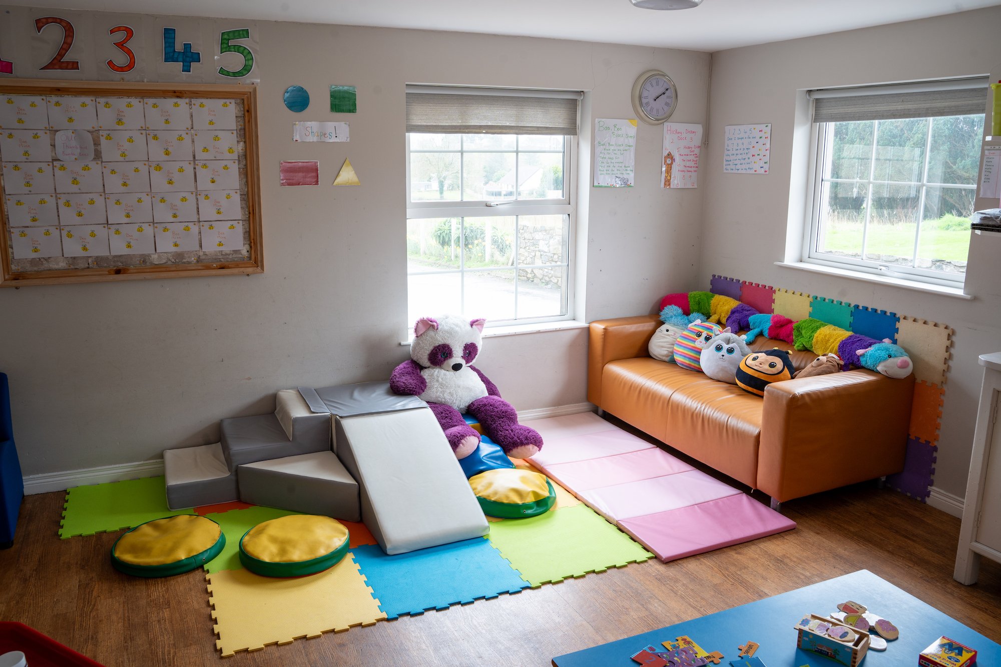 A cozy playroom corner with colorful foam mats, stuffed animal toys including a panda, and a small orange sofa with plush cushions, situated near windows with a view of a grassy outdoor area.