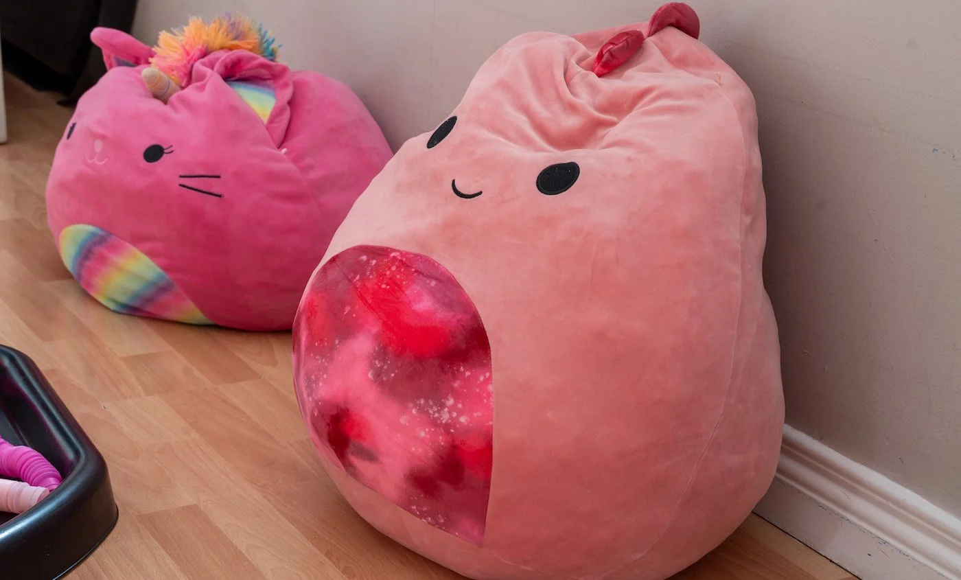 Two plush stuffed animals resembling strawberries with cute faces, placed on a wooden floor against a wall. The one in the foreground has a galaxy-themed heart-shaped detail on its tummy.