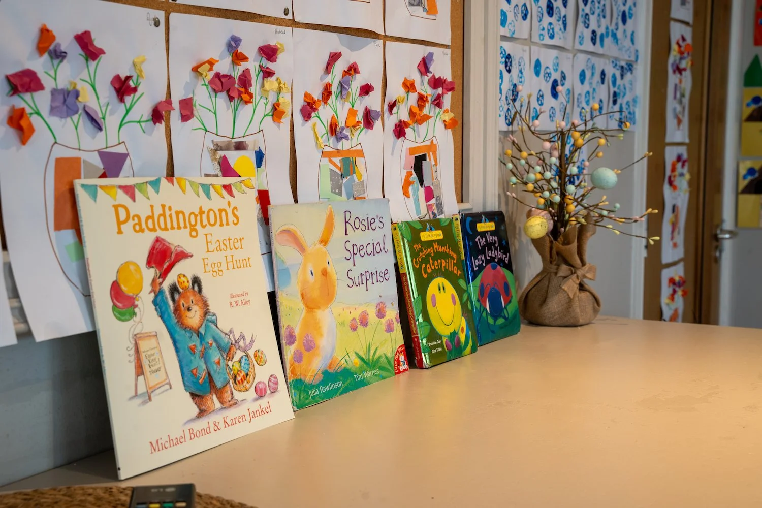 Children's books with Easter and spring themes, colorful paper flower artwork on the wall, a decorative branch with pastel eggs in a cloth bag, in a classroom or library setting.