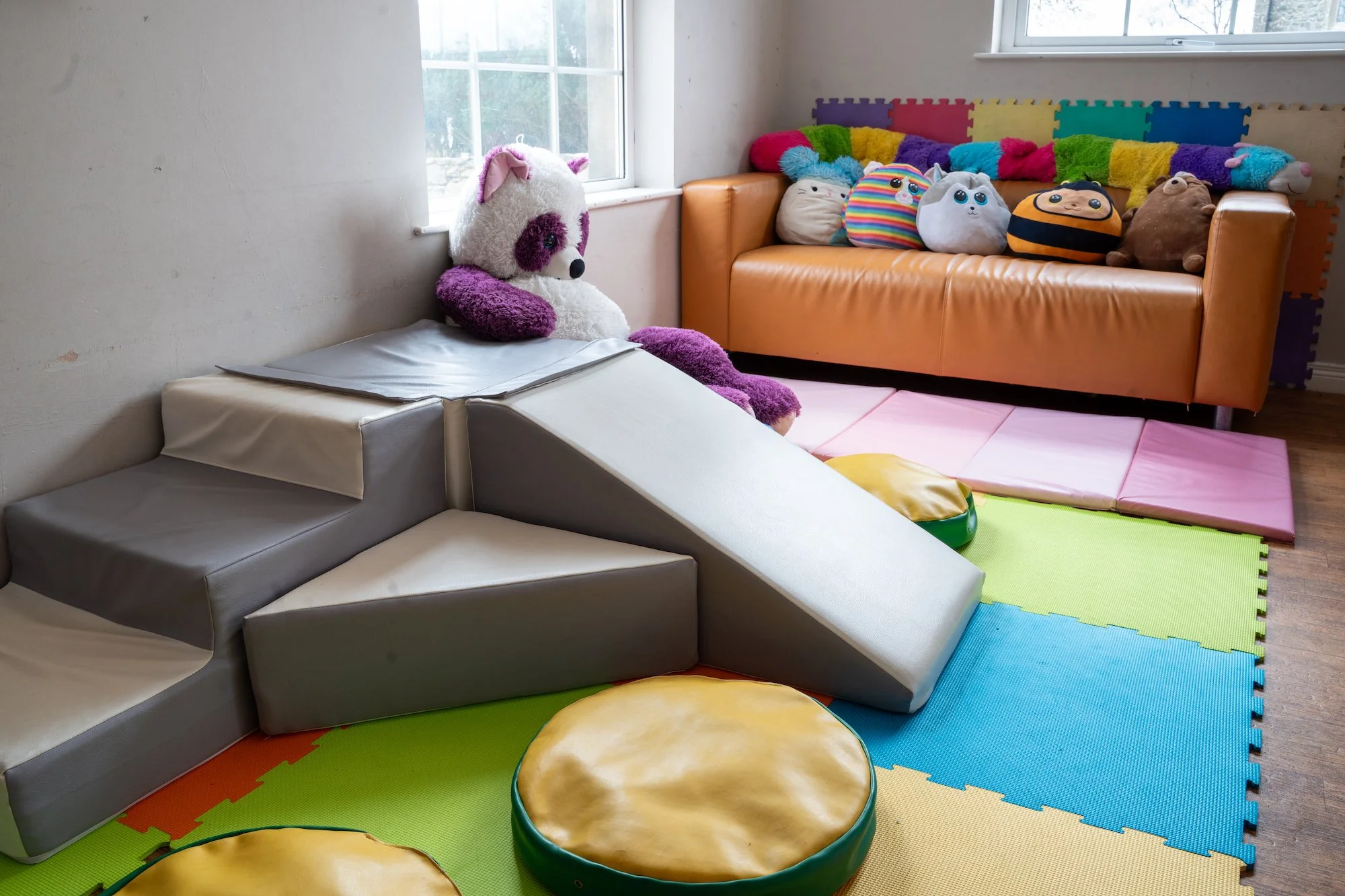Indoor playroom with colorful foam mats, plush toys, and a small couch decorated with pillow plush toys.