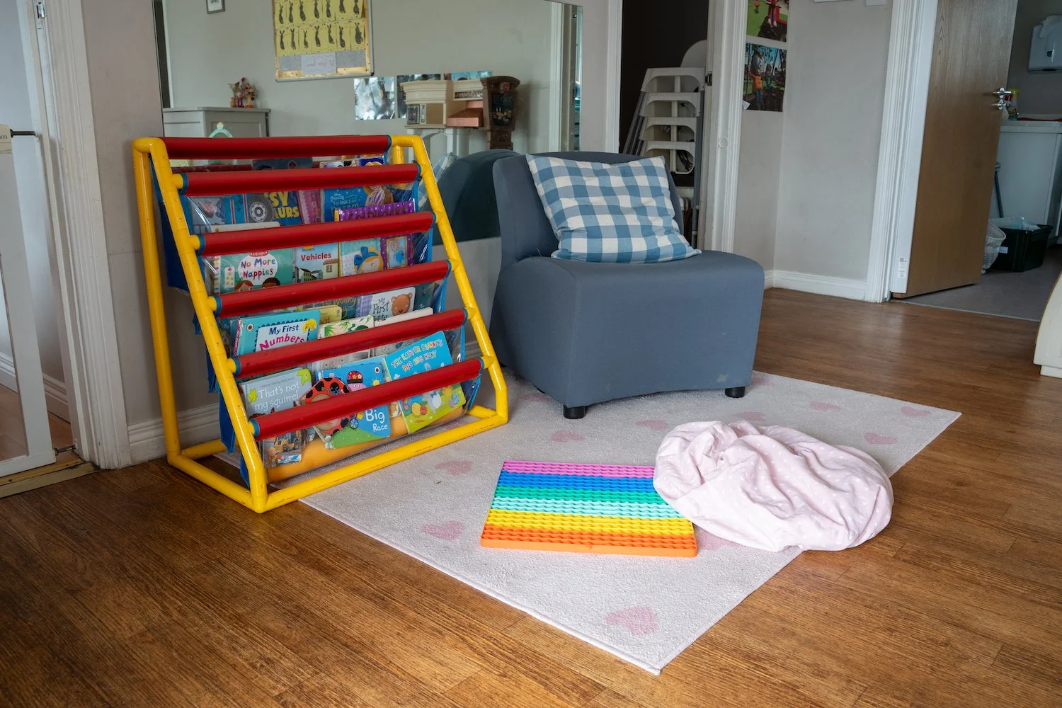 Child's playroom with a book rack filled with colorful books, a gray armchair with a checkered pillow, a multicolored sensory pad, and a soft pink blanket on the floor, on a white rug with pink heart patterns in front of wooden flooring.