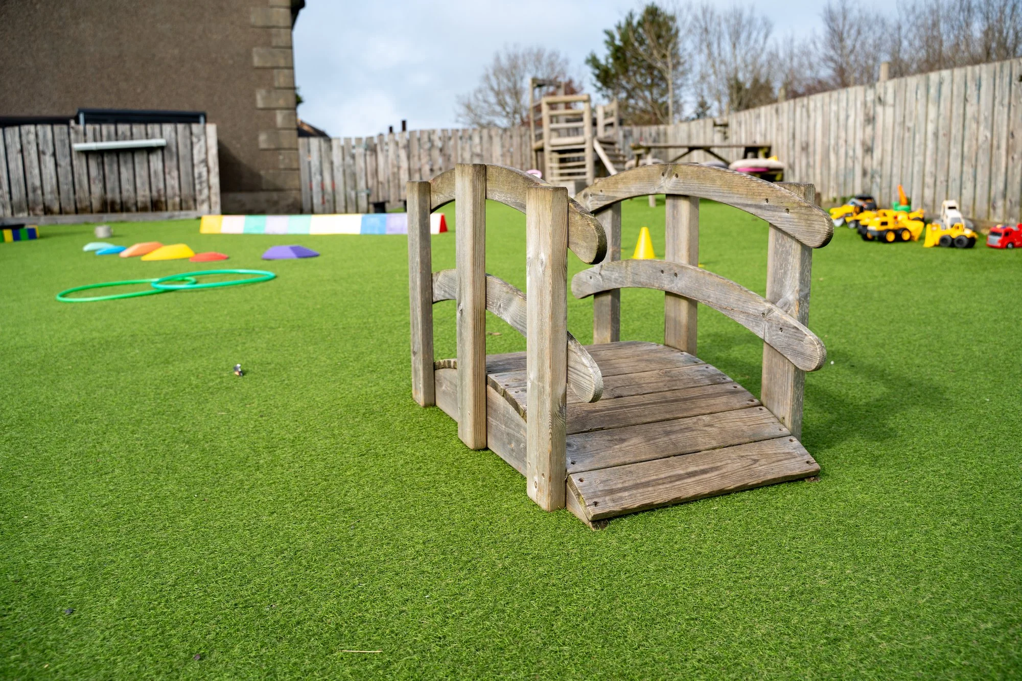 A small wooden bridge in a children's playground on a green artificial turf surrounded by various toys and equipment, with a wooden fence and houses in the background.