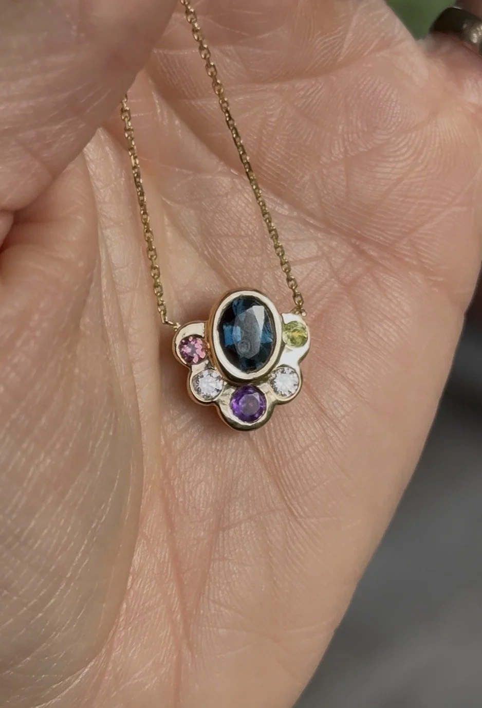 Collier avec pendentif contenant un grand pierre noire centrale entourée de plusieurs petites pierres colorées, porté sur la main d'une personne.