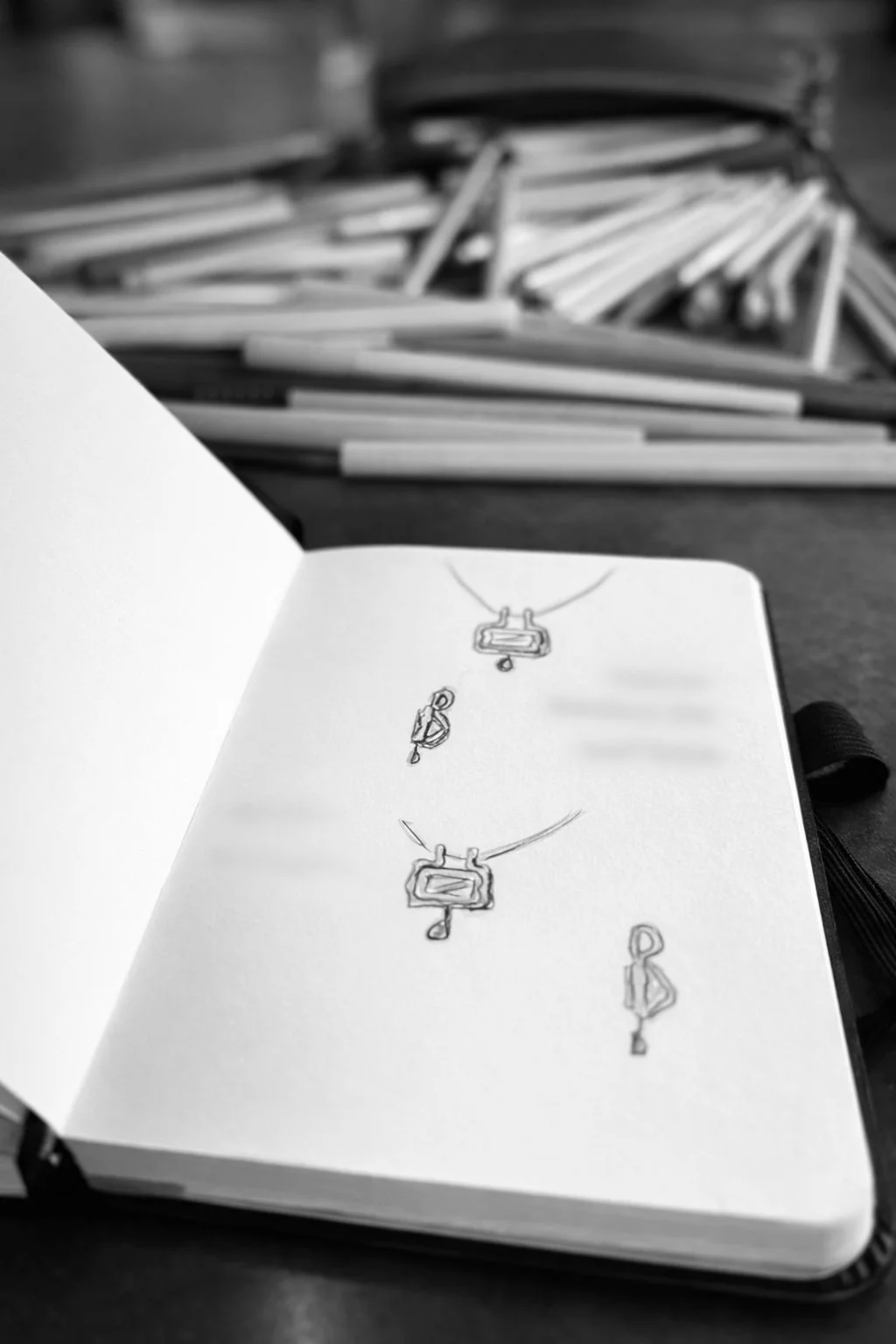 Un carnet ouvert avec des croquis de colliers et pendentifs, posé sur une table avec des crayons en arrière-plan.