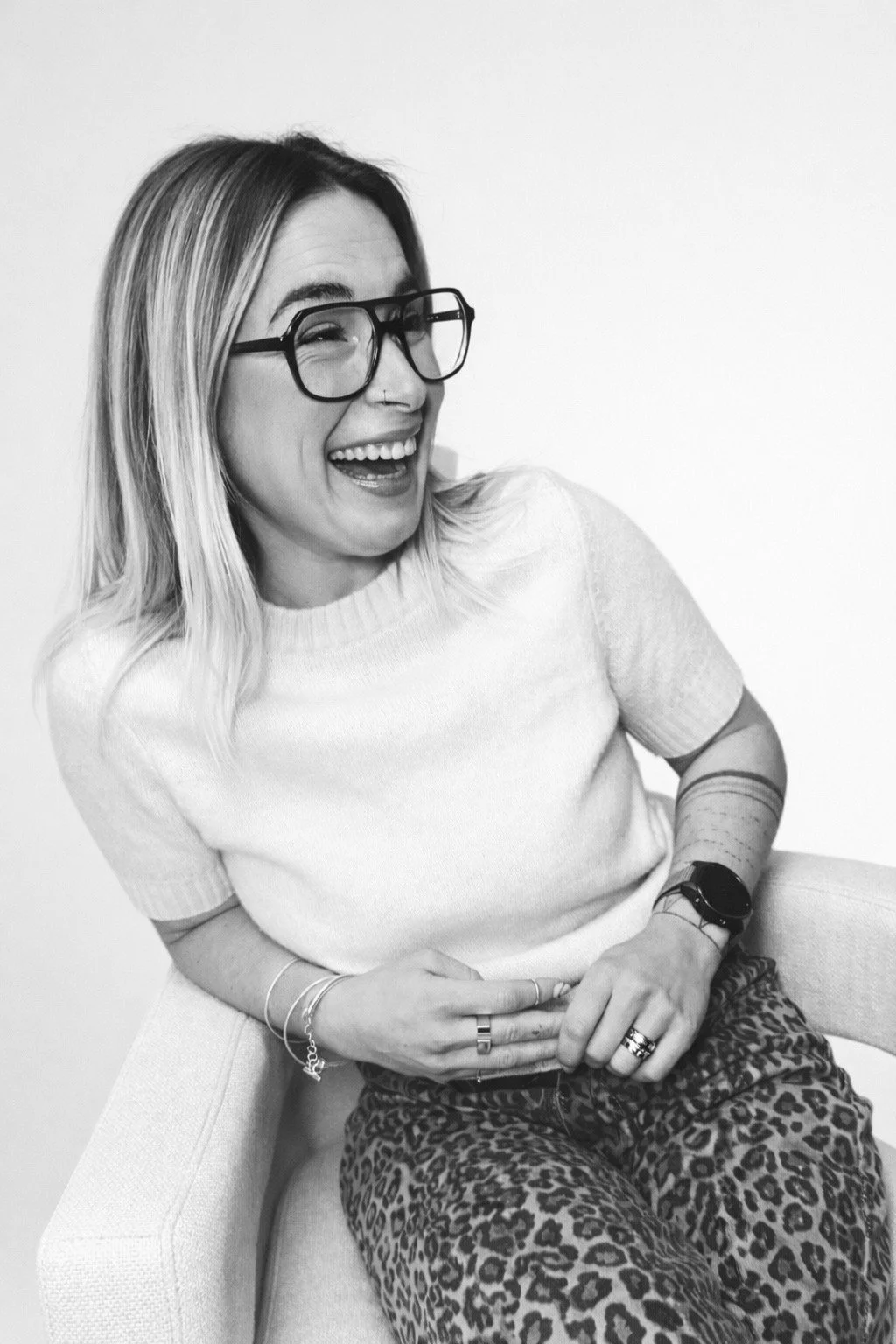 Une femme souriante avec des lunettes, portant un pull clair et un pantalon à motif léopard, assise sur une chaise blanche contre un fond blanc.