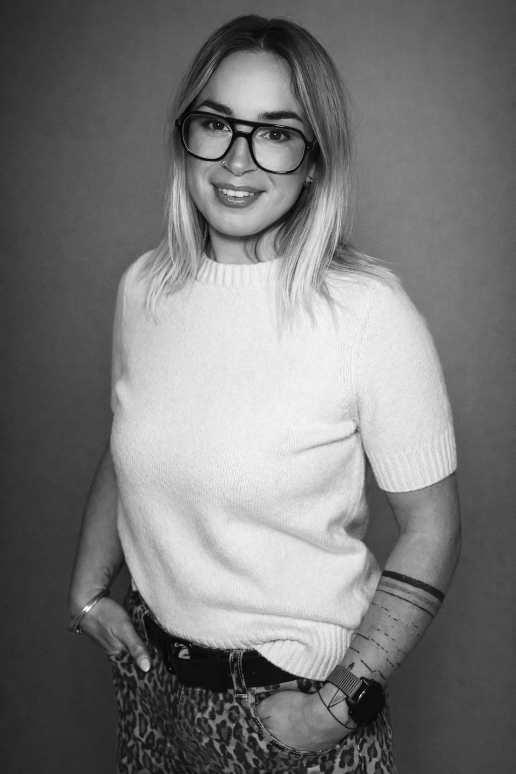 Portrait en noir et blanc d'une femme aux cheveux blonds, portant des lunettes, un pull blanc côtelé, un pantalon léopard, et une montre, souriante, avec un fond uni.