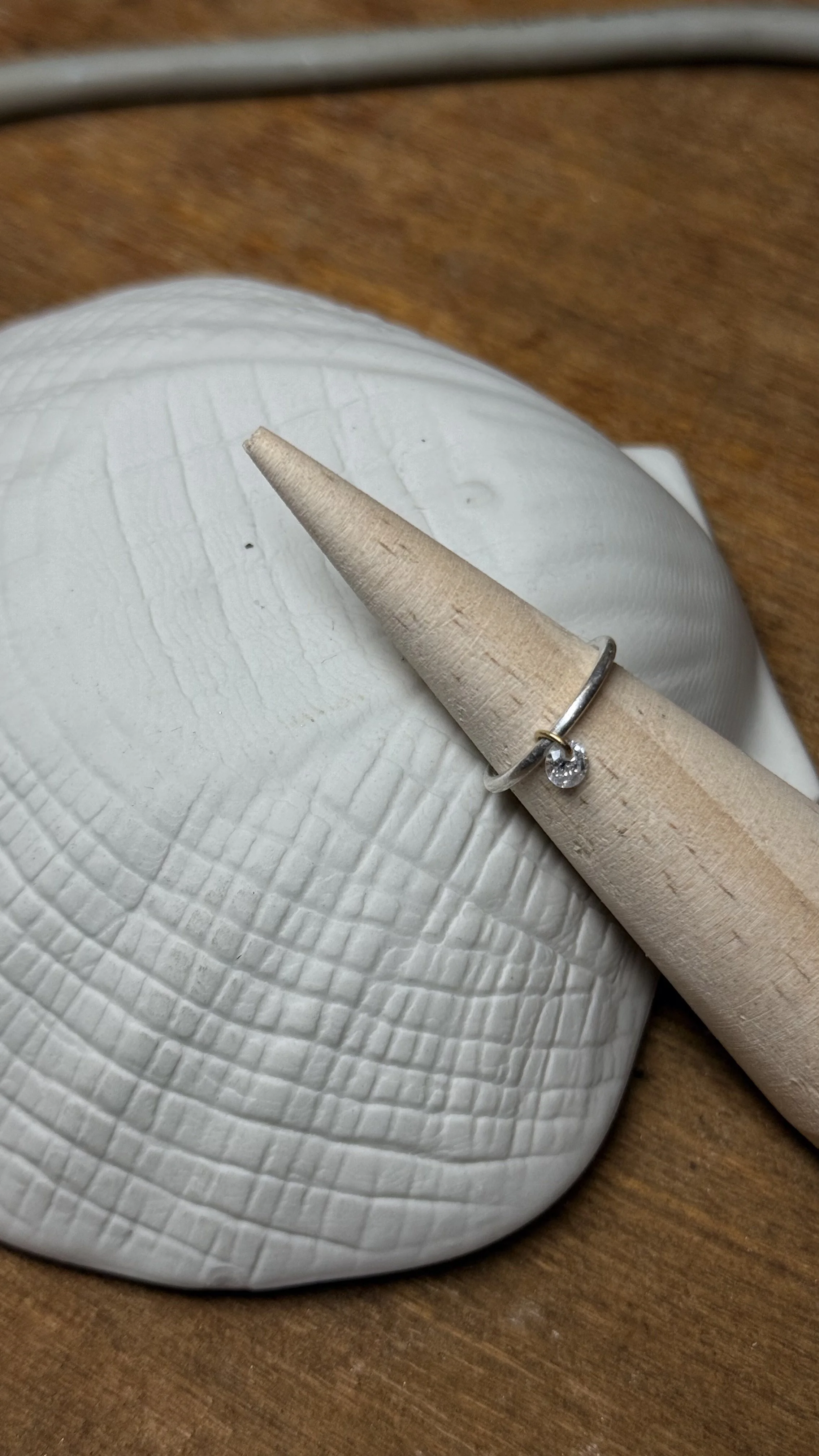 Une bague avec un diamant est posée sur un anneau en bois, le tout repose sur une surface en céramique blanche avec une texture rayée.