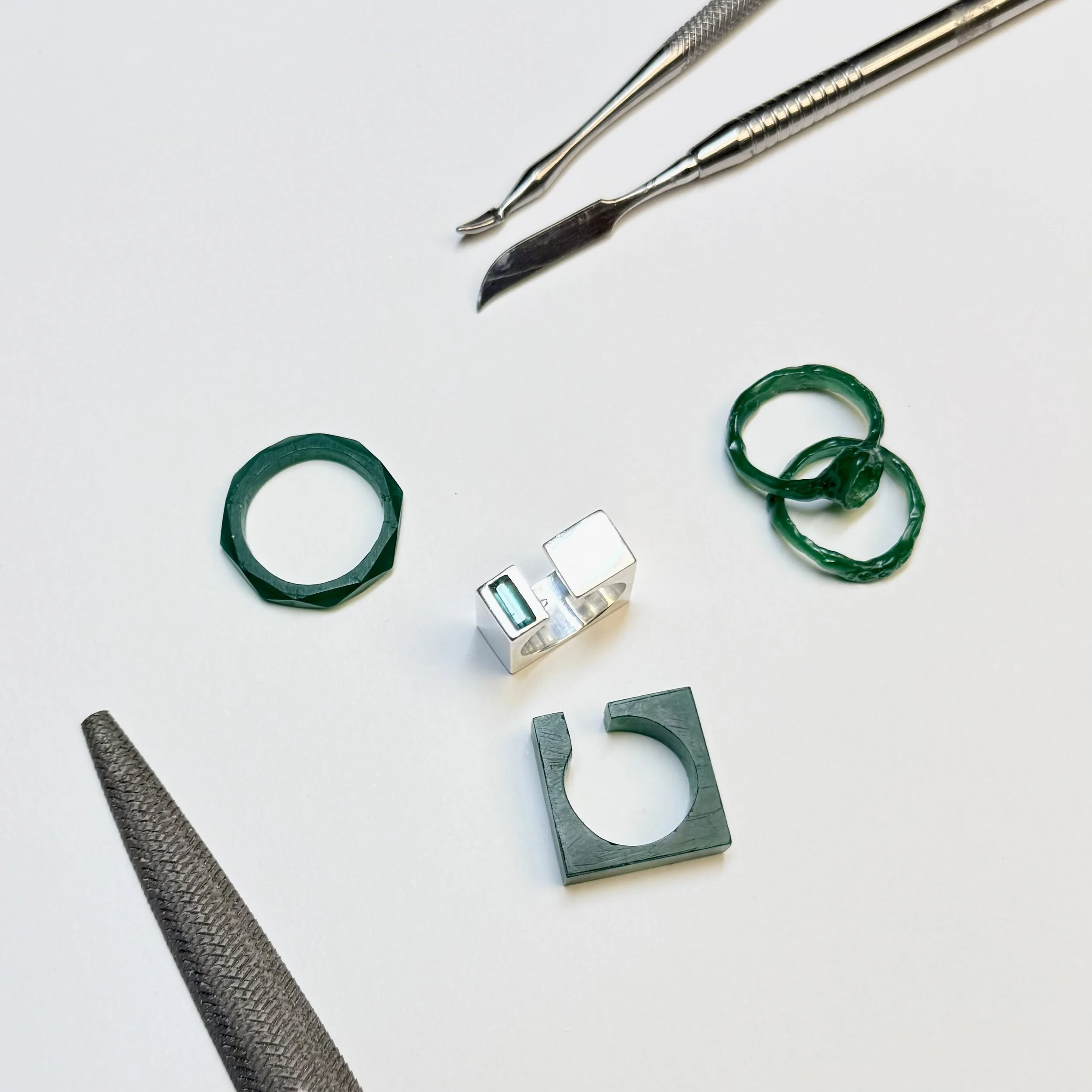 Une collection d'outils et d'objets métalliques, notamment un crochet en métal, une poignée en caoutchouc, une petite boîte métallique, et quelques anneaux en plastique vert, disposés sur une surface blanche.