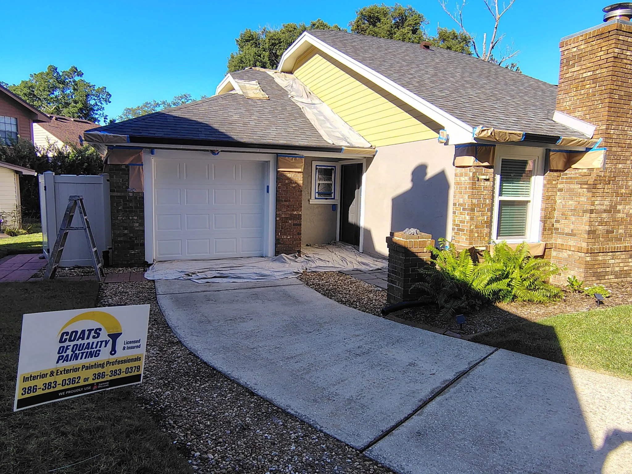Full front view of an Orlando home undergoing exterior painting, showing a garage, brick chimney, yellow gable, protected entry, and Coats of Quality Painting yard sign.