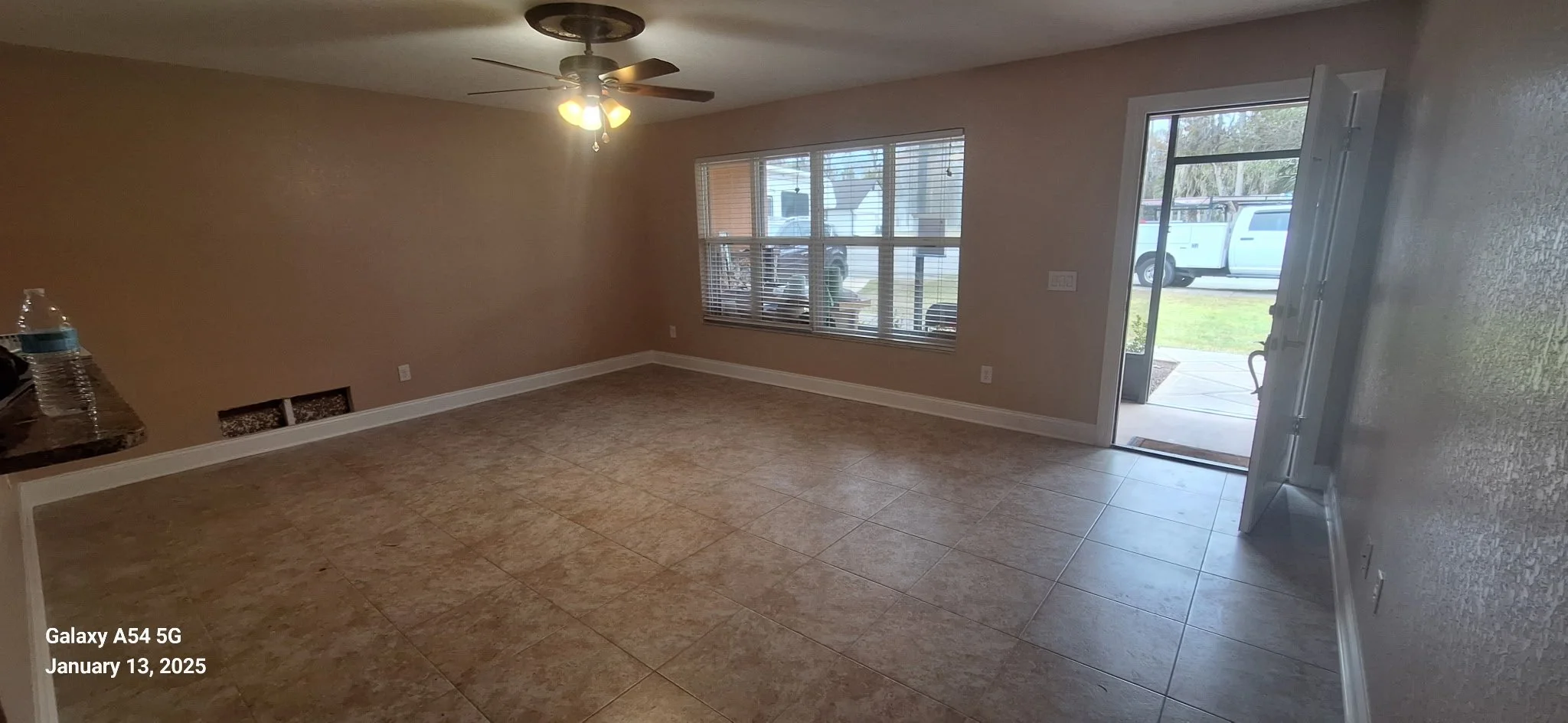 Empty living room with beige walls, large window with blinds, ceiling fan with lights, and tile flooring. An open door leads outside to a grassy yard and parked vehicles.
