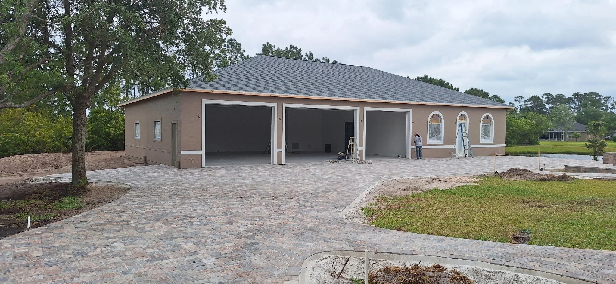 Construction site of a residential house with a paved driveway, two large open garage bays, a worker, ladders, and new landscaping.
