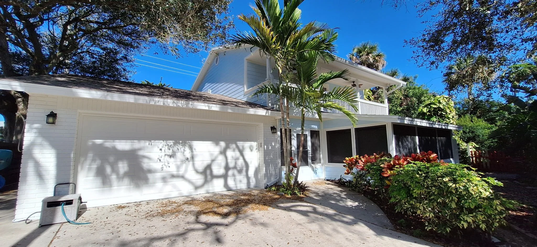 A white two-story house with a screened porch and balcony, surrounded by lush green plants and tall trees under a clear blue sky.
