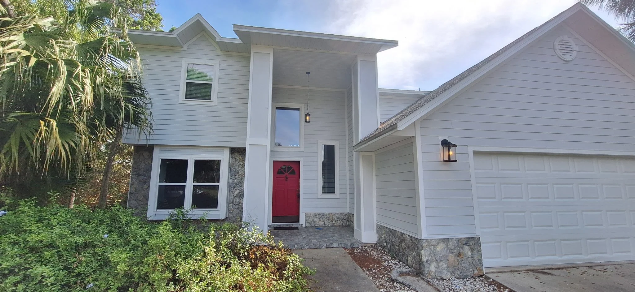 Modern two-story house with white siding, a stone foundation, red front door, and a garage with a White door. There are black exterior lights and a hanging porch light, surrounded by greenery and palm trees.