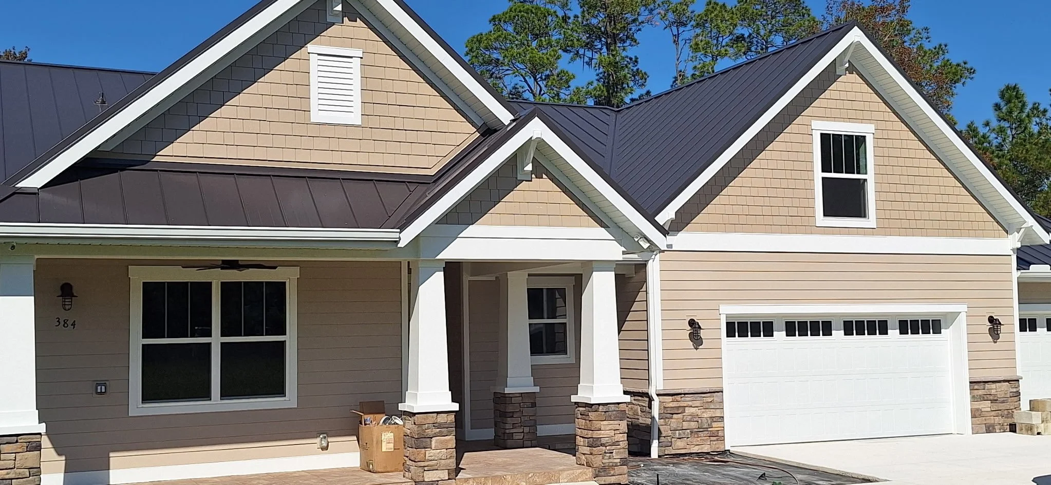 Front view of a modern two-story house with beige siding, stone accents, and a black metal roof. There are large windows, a small entrance porch with white columns, and a white garage door. Green trees are visible in the background.