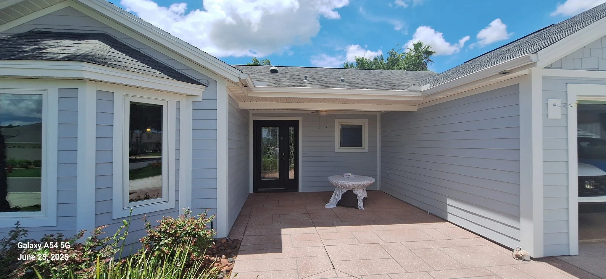 Exterior view of a house with light gray siding, large windows, a black front door with glass, and a covered patio area. The patio has a tiled floor and a small round table covered with a cloth. There are some plants near the corner of the house and a partly cloudy sky above.