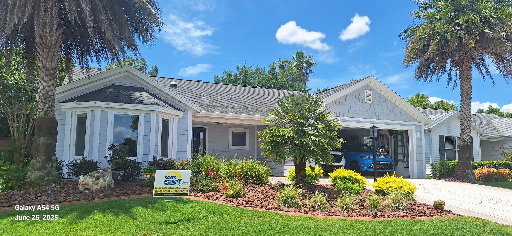 Front yard view of a house with white siding, two garage doors with a blue car parked inside, a landscaped garden with palm trees, bushes, and flowers, a lamp post, and a driveway. There is a sign for a painting company in the garden, and the sky is blue with some clouds. The photo is dated June 25, 2025, taken with a Galaxy A54 5G.