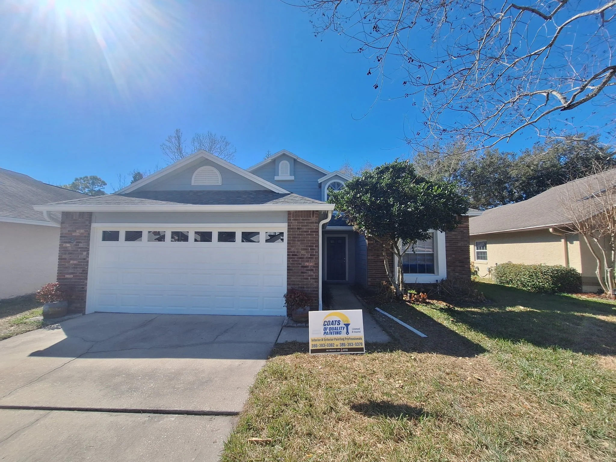 A suburban house with a white garage door, brick accents, and a covered front porch. There are trees and a lawn in front, with a sign for painting services on the lawn.