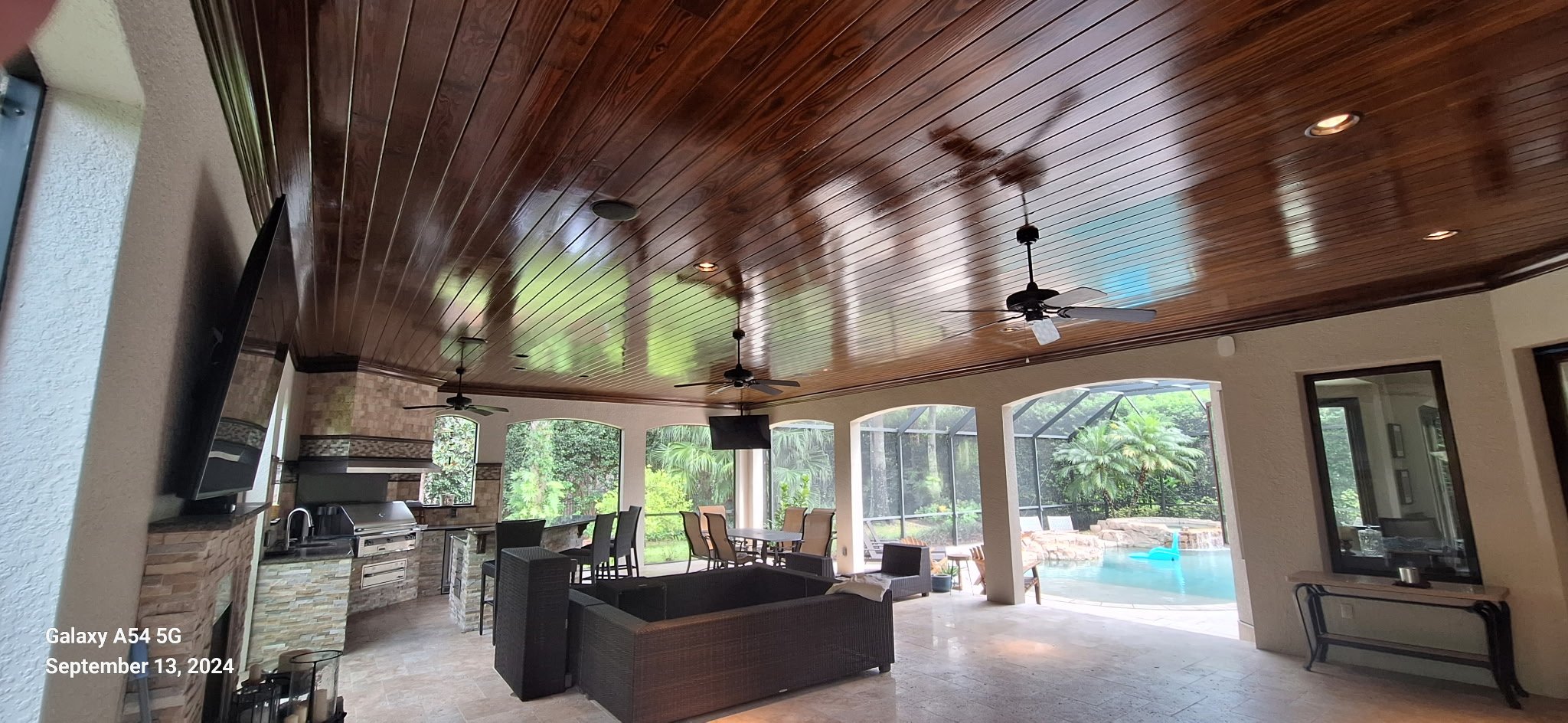 Indoor patio area with a shiny wooden ceiling, ceiling fans, a wall-mounted TV, a stone fireplace with a grill, dining tables and chairs. Includes a view of a backyard swimming pool with a floating blue pool toy and lush greenery outside.