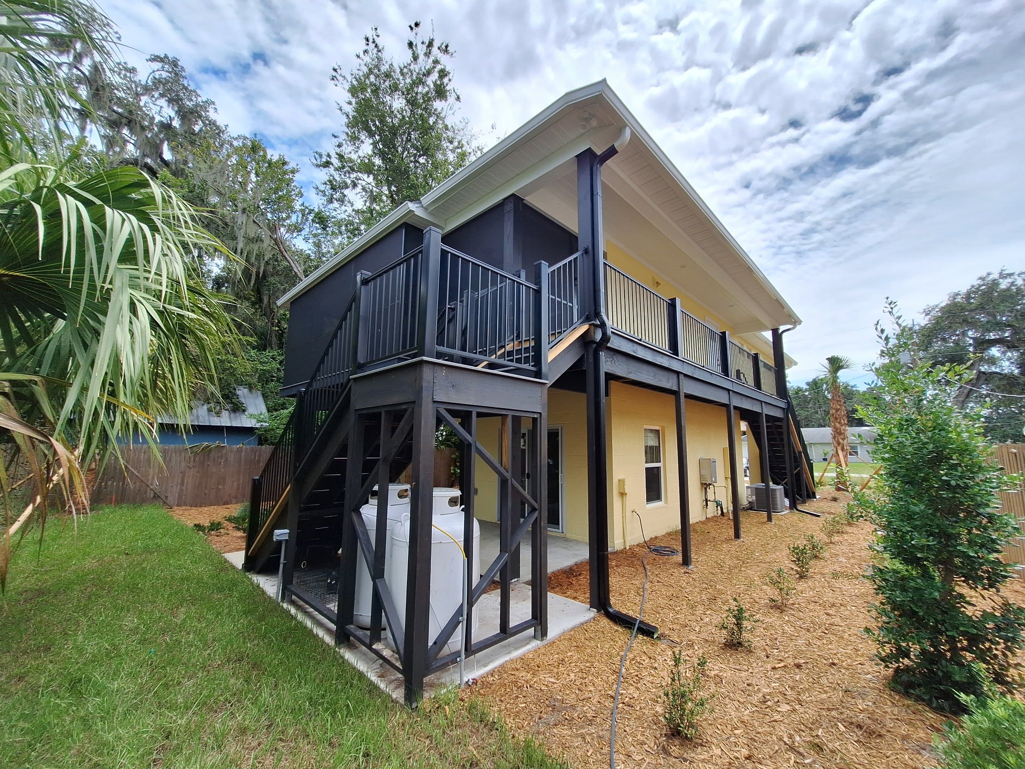 Two-story house with a yellow exterior, black railings, and black stairs leading to the upper deck, surrounded by trees and greenery.