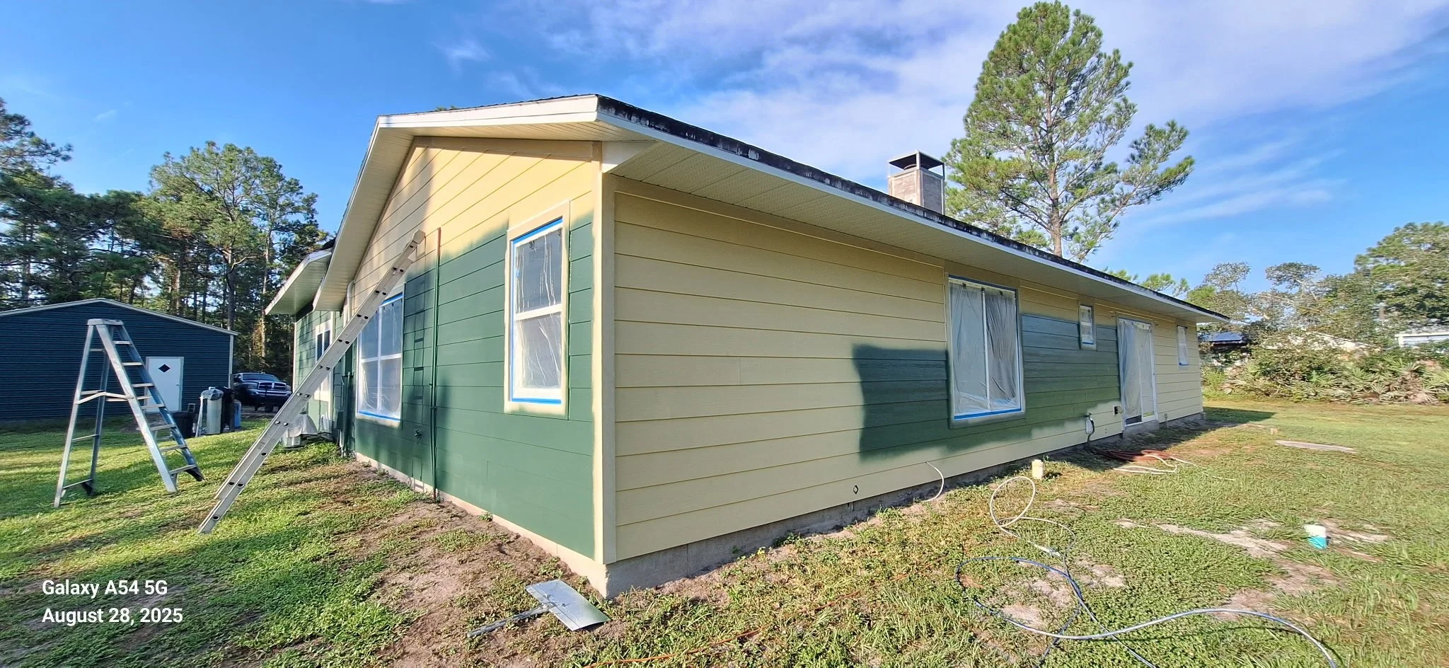 Side view of a New Smyrna Beach home during an exterior painting project, highlighting professional preparation of Hardie board siding. This stage shows the careful transition from the original yellow exterior to a modern green color designed to with