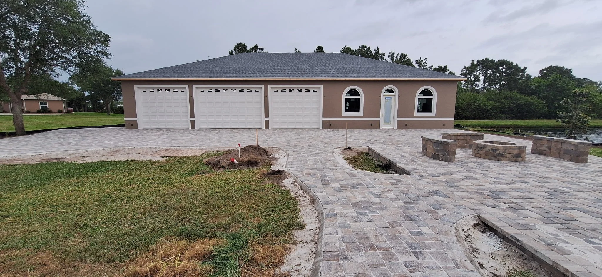 Port Orange FL exterior house painting project featuring freshly painted tan stucco home with white trim, three-car garage, and newly installed paver driveway and patio overlooking landscaped yard.
