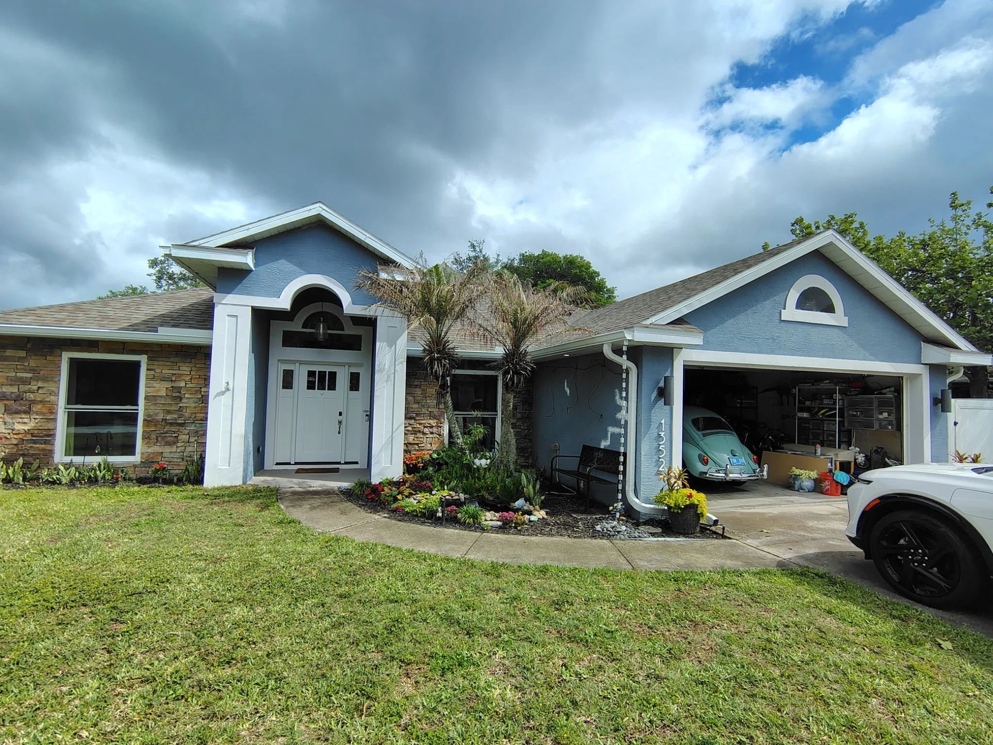 A blue house with a stone facade and white trim, a front yard with a lawn and flower bed, a driveway with a classic Volkswagen Beetle in the garage, and a white car parked outside. The cloudy sky is overhead.