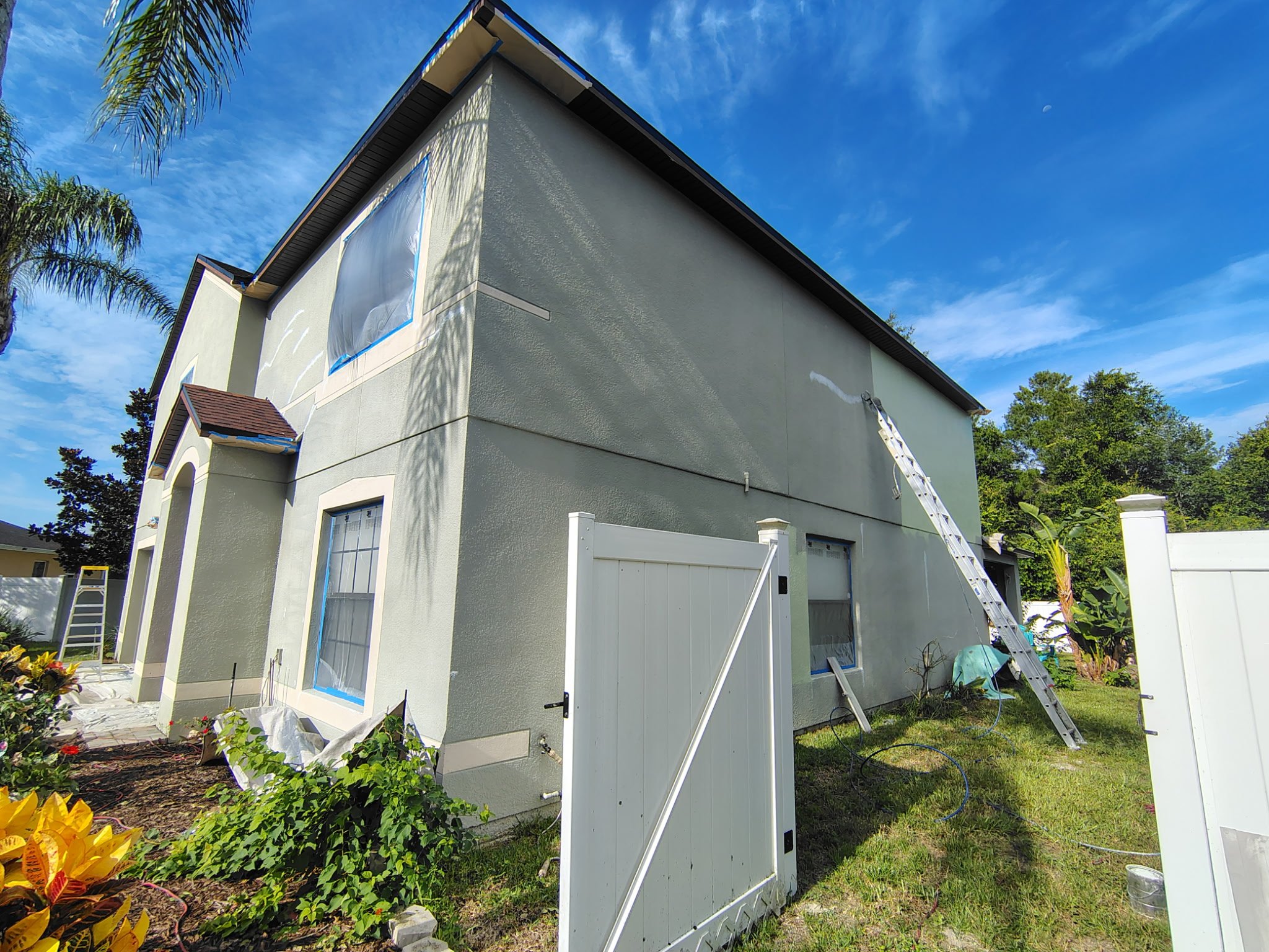 Side of two-story Deltona stucco home during exterior painting with masked windows, ladder and prepared wall surface.