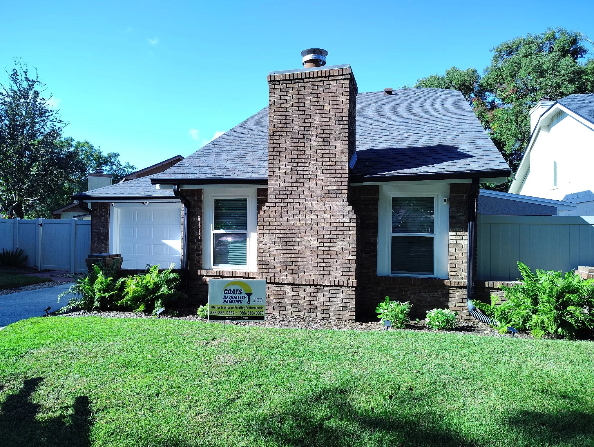 Side of an Orlando house after exterior painting with light neutral gable siding, white fascia, natural brick chimney, and white privacy fence.