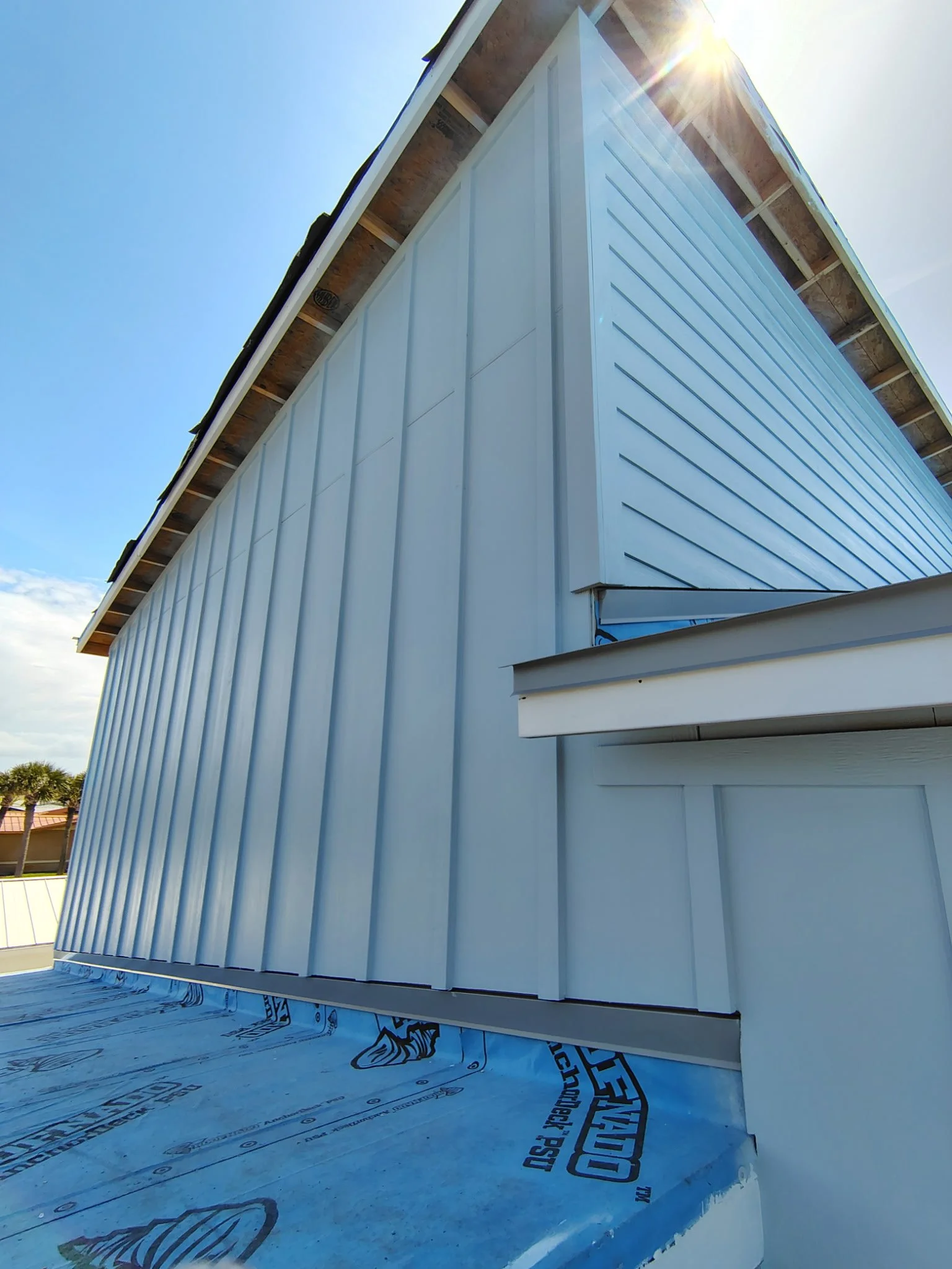 Close-up of a building under construction with white siding and a metal roof, sunlight shining from the top right corner.