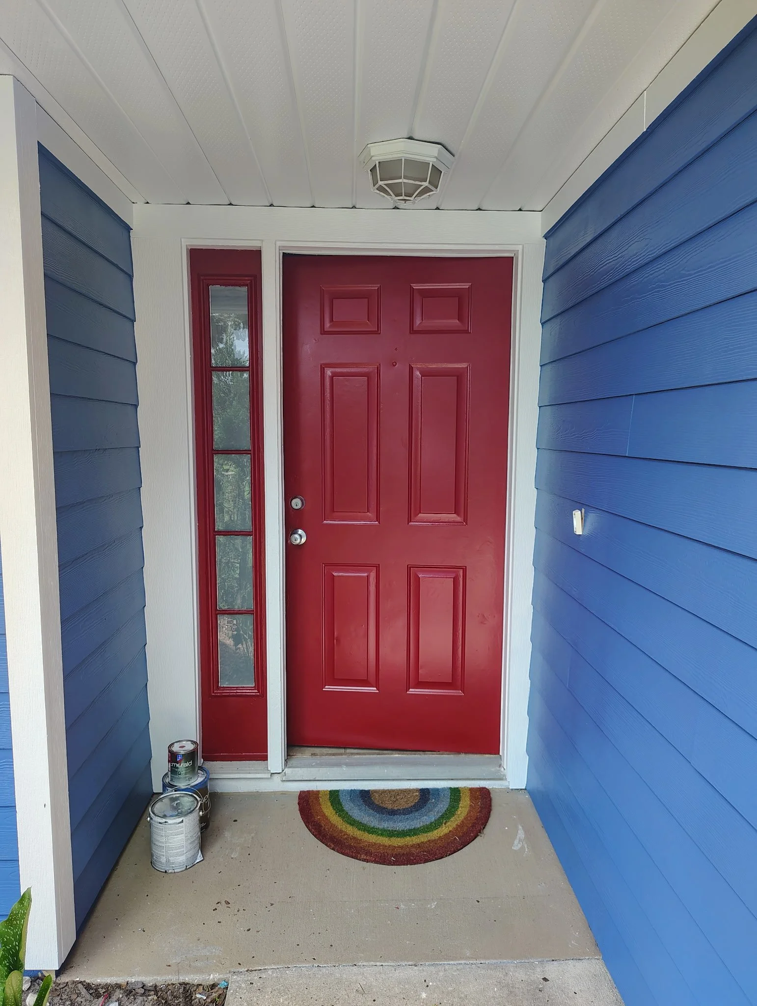The home's completed entrance now features a rich red front door and sidelight surrounded by bright white trim and blue siding. The contrasting entry creates a strong focal point within the new exterior color palette.