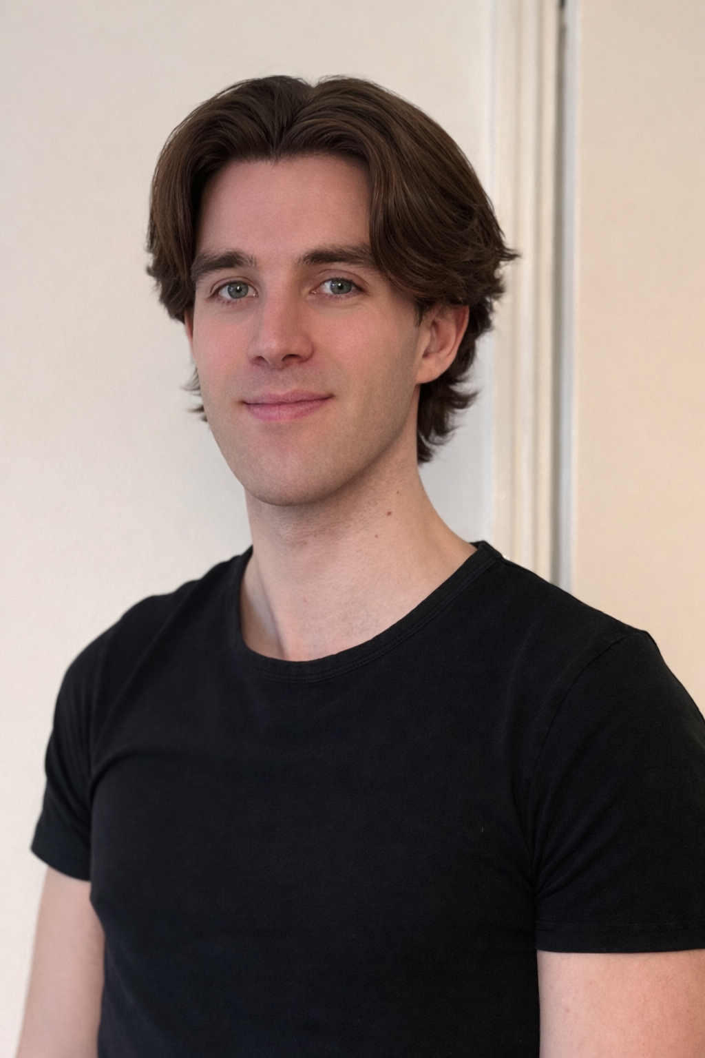 A young man with shoulder-length brown hair, fair skin, and blue eyes, wearing a black T-shirt, standing against a plain light-colored wall.