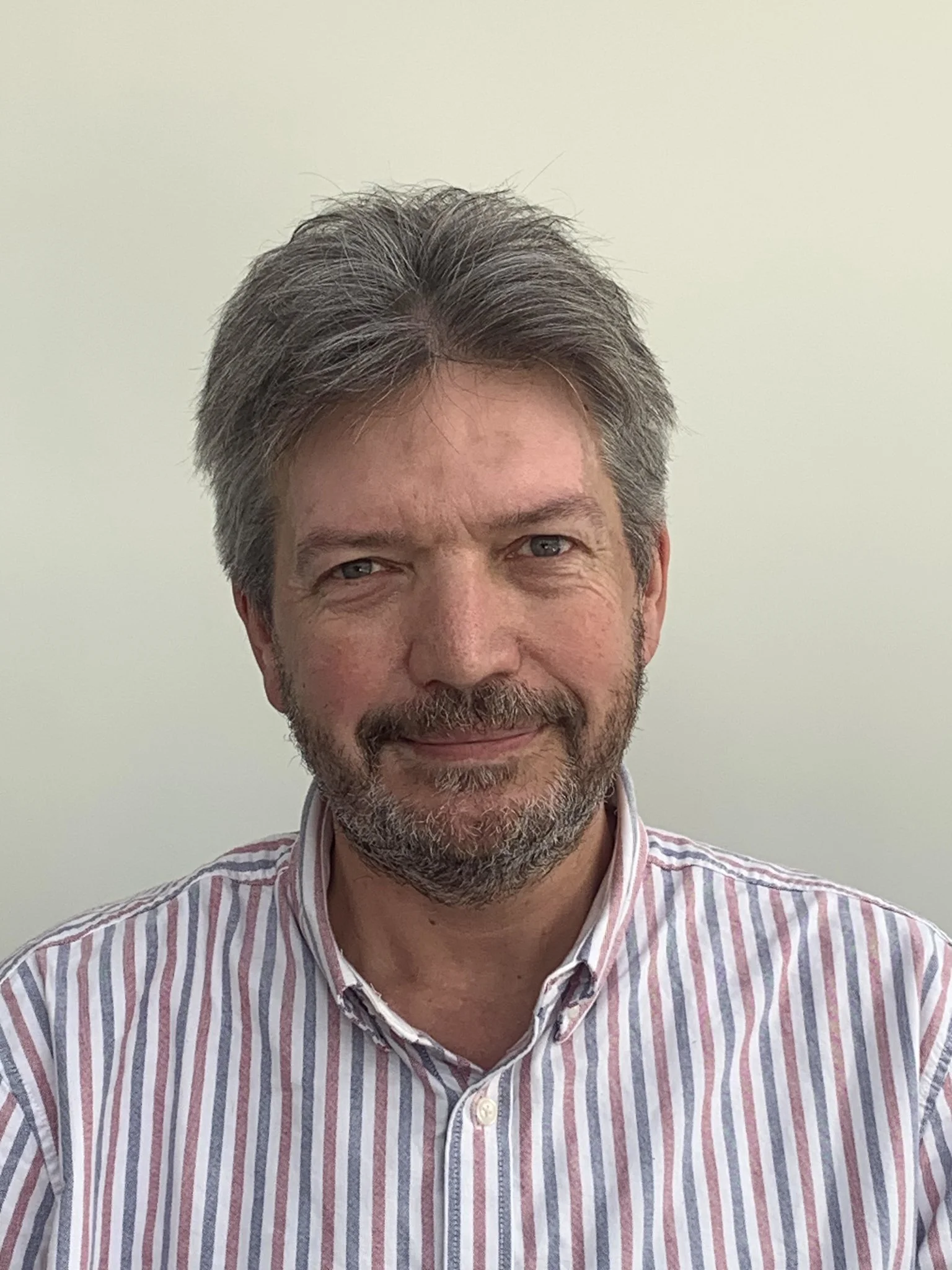 A middle-aged man with salt-and-pepper hair and beard, smiling, wearing a striped button-down shirt, standing against a plain light-colored background.