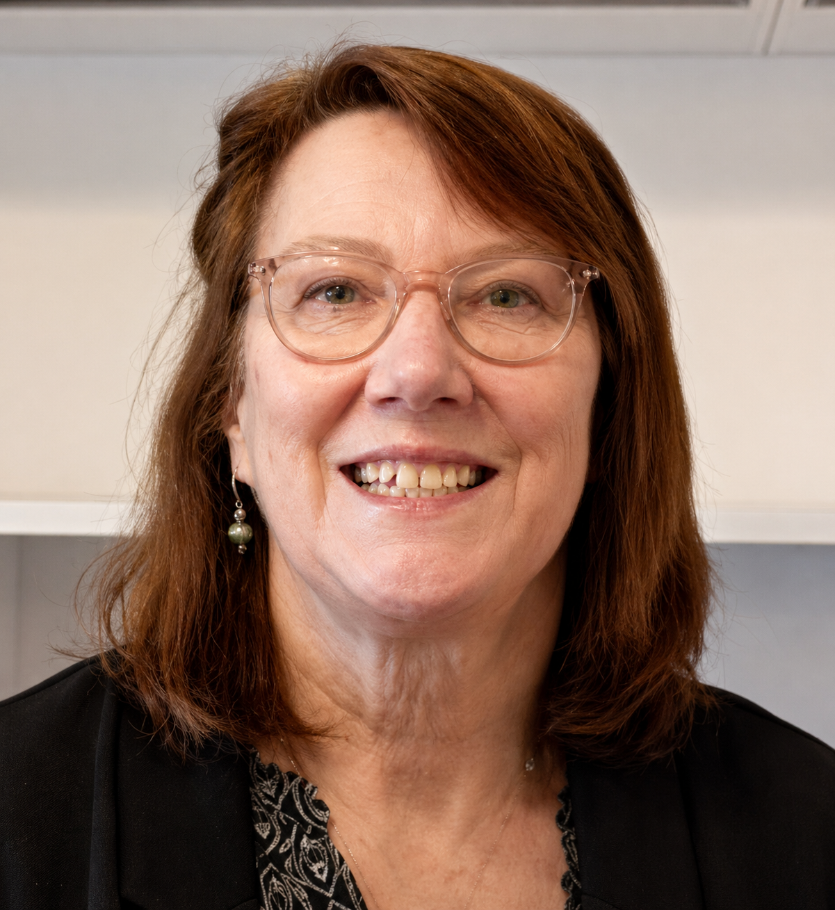 Close-up of a smiling woman with red hair, glasses, and earrings, wearing a black jacket and patterned blouse, in an indoor setting.