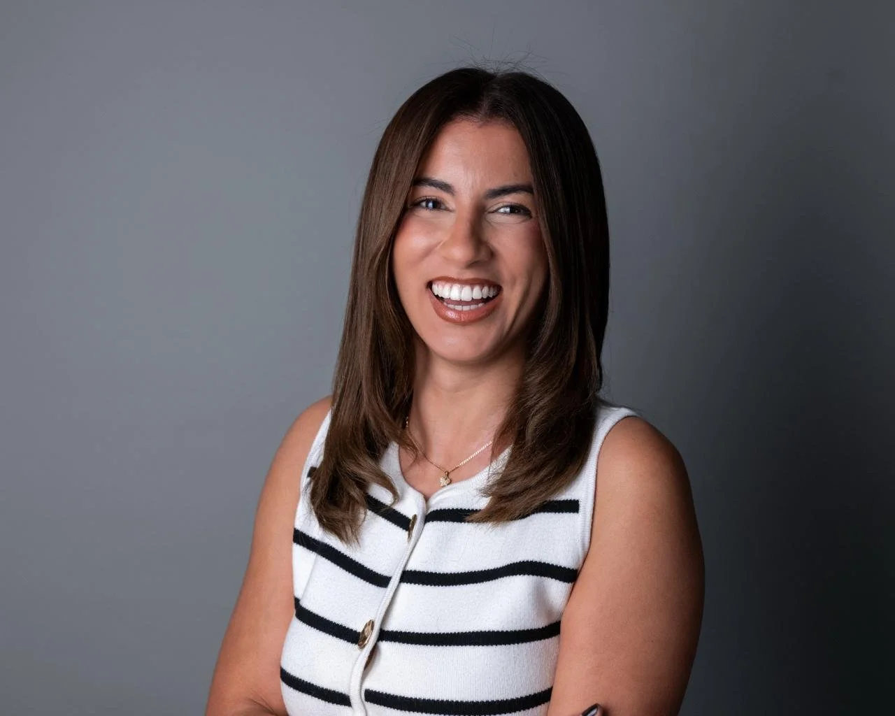 Smiling woman with shoulder-length brown hair wearing a sleeveless white top with black horizontal stripes against a gray background.
