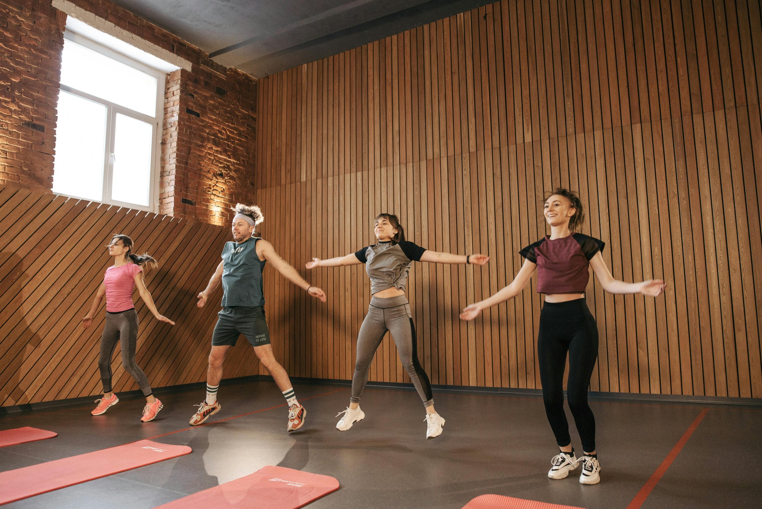 Four people jumping during an indoor workout or dance class in a room with wooden wall panels and a large window.