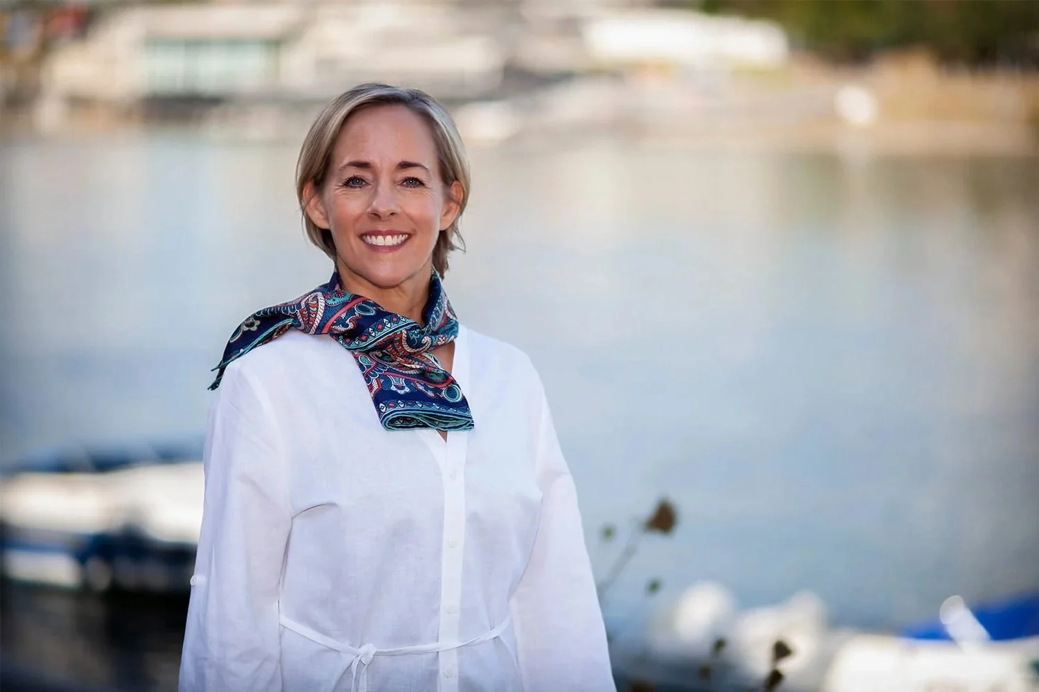 Julie Morrow woman outdoors near a body of water wearing a white shirt and a colorful patterned scarf.