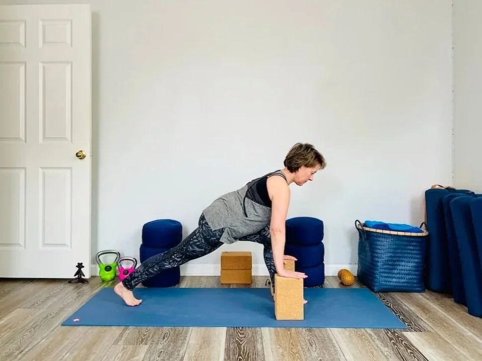 Julie Morrow  practicing yoga in a room with fitness equipment, in a plank position with hands on yoga blocks, wearing workout clothes, on a blue yoga mat.