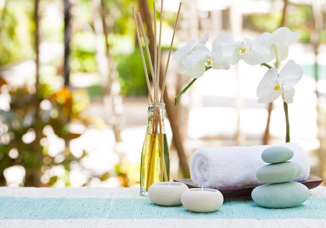 Spa setting featuring a tall glass vase with white orchids and reed diffuser, a rolled white towel, smooth stacked stones, and small ceramic candle holders on a light blue and white cloth with a blurred outdoor background.