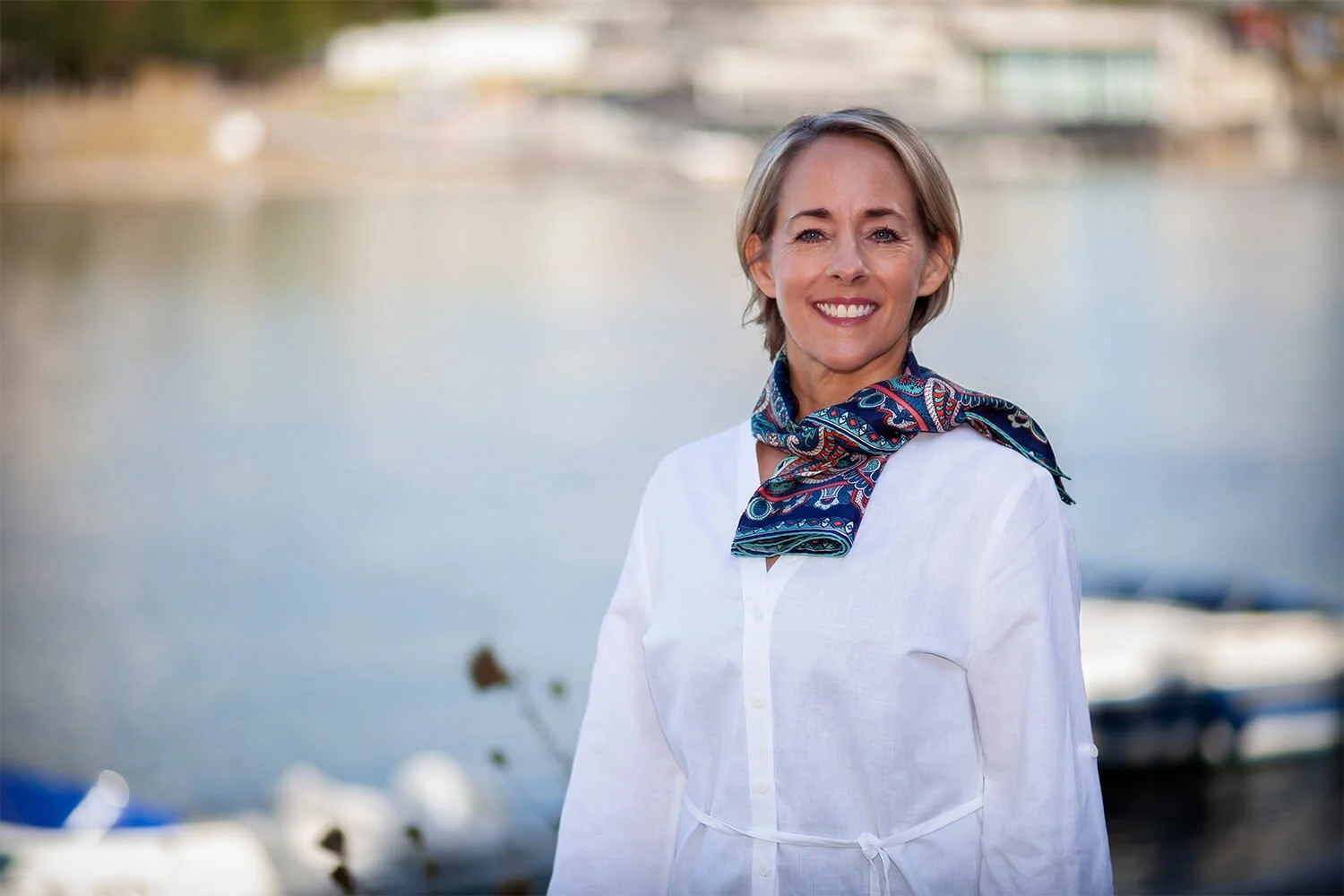 Julie Morrow smiling outdoors near a body of water, wearing a white shirt and a blue patterned scarf.