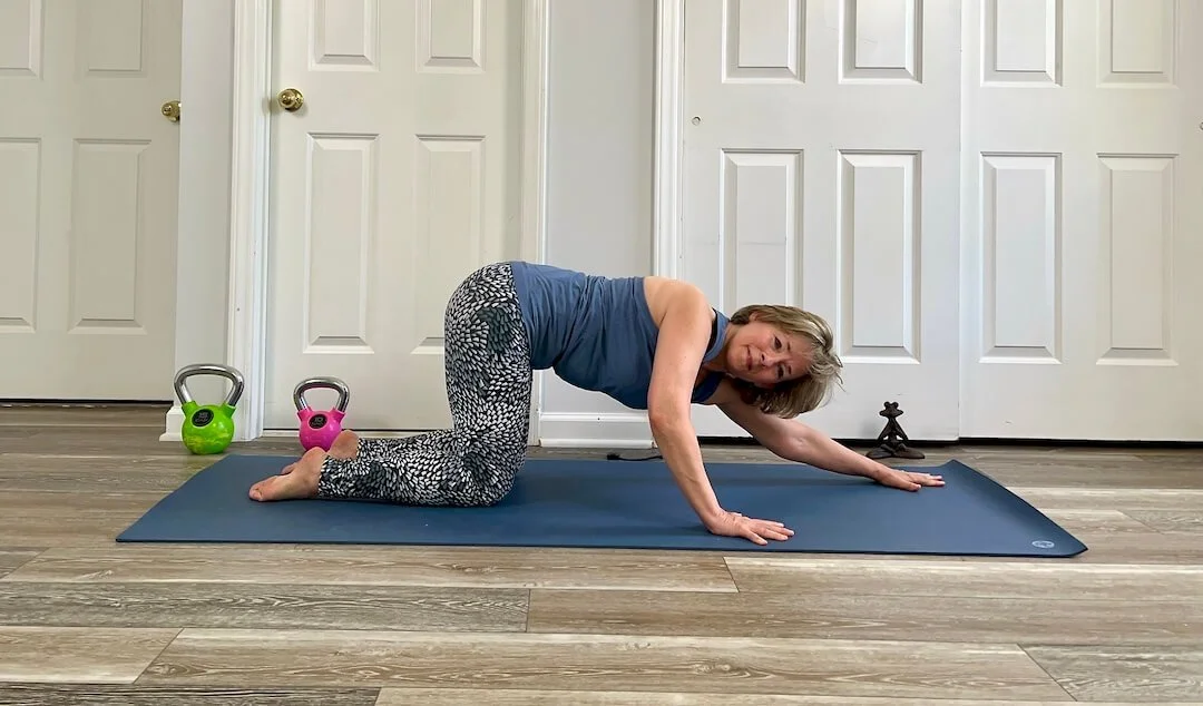 Julie Morrow performing a yoga pose on a blue exercise mat in front of closed white closet doors. She is on her hands and knees, with her right arm extended forward and her face turned towards the camera.