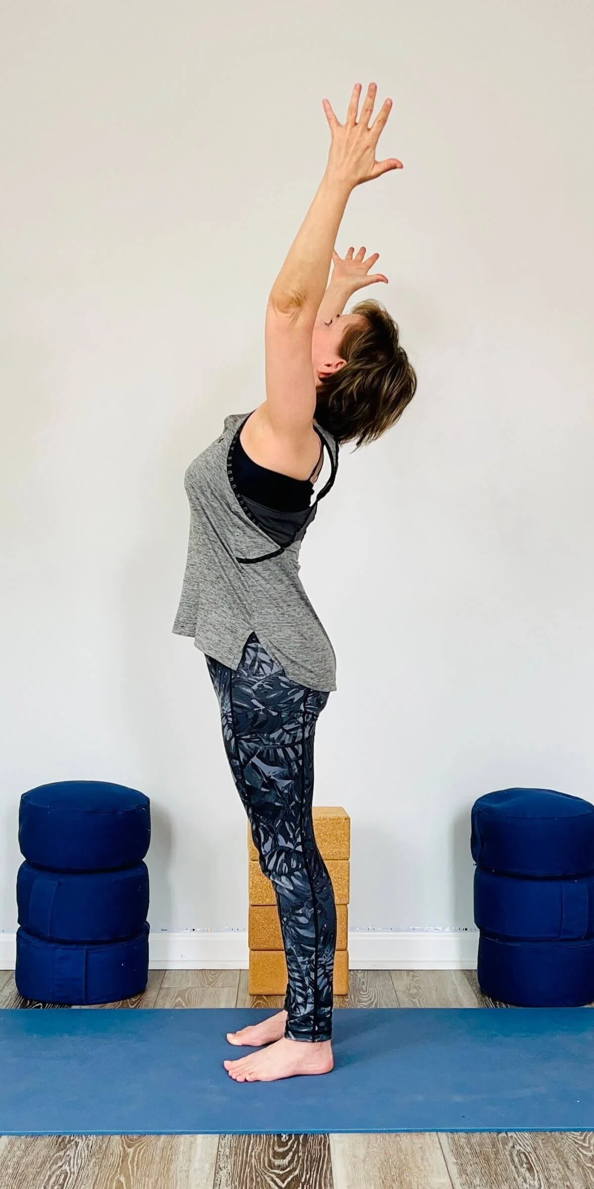 Julie Morrow performs a yoga pose with arms extended upward, head tilted back, eyes closed, standing on a yoga mat in a studio with pillows and wooden blocks in the background.
